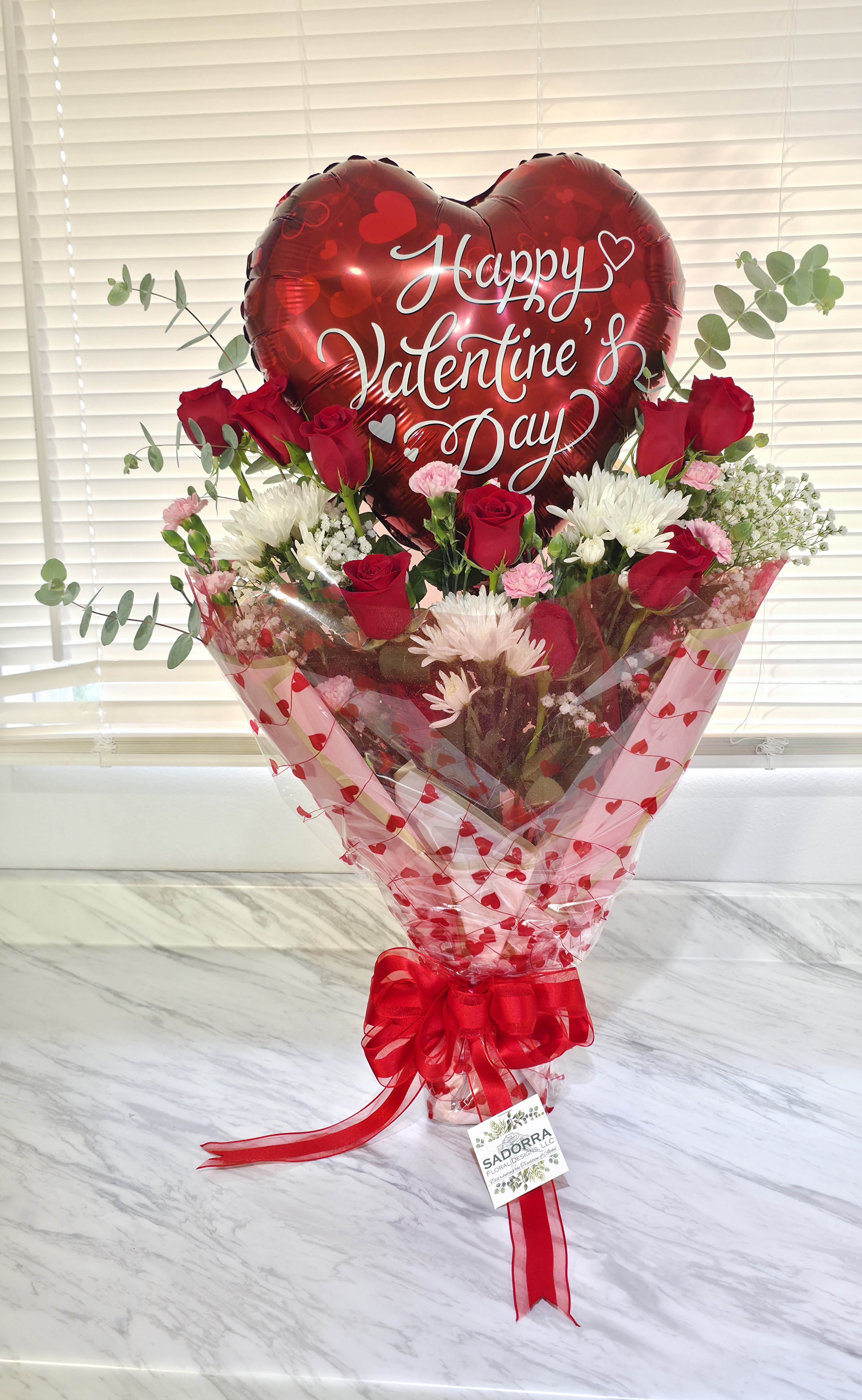"Be My Valentine's" Arrangement  - "Be My Valentine's Arrangement." Is an array of 12 Red Designers Choice Roses, pink mini carnations, baby's breath and greenery based in a watered oasis and wrapped in gift paper &amp; a "Happy Valentine's" Mylar Balloon! Made for that special Valentine's! 