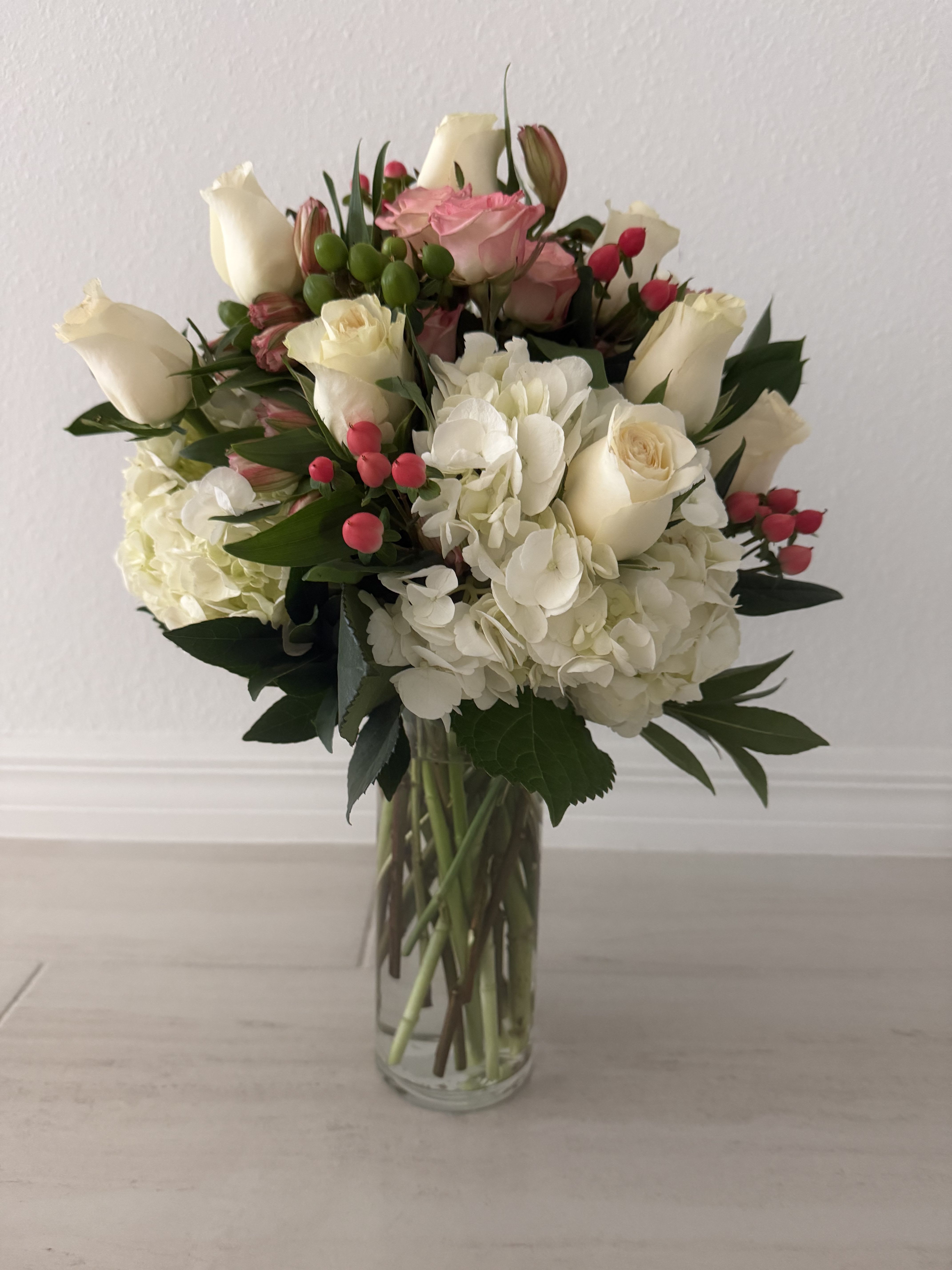 Blushing Elegance Bouquet - An elegant floral arrangement featuring white hydrangeas beautifully paired with white and soft pink roses, accented with fresh greenery and delicate seasonal berries. Designed in a clear glass vase, this bouquet expresses grace, purity, and gentle romance. Perfect for anniversaries, birthdays, sympathy gestures, or to send a message of love and appreciation