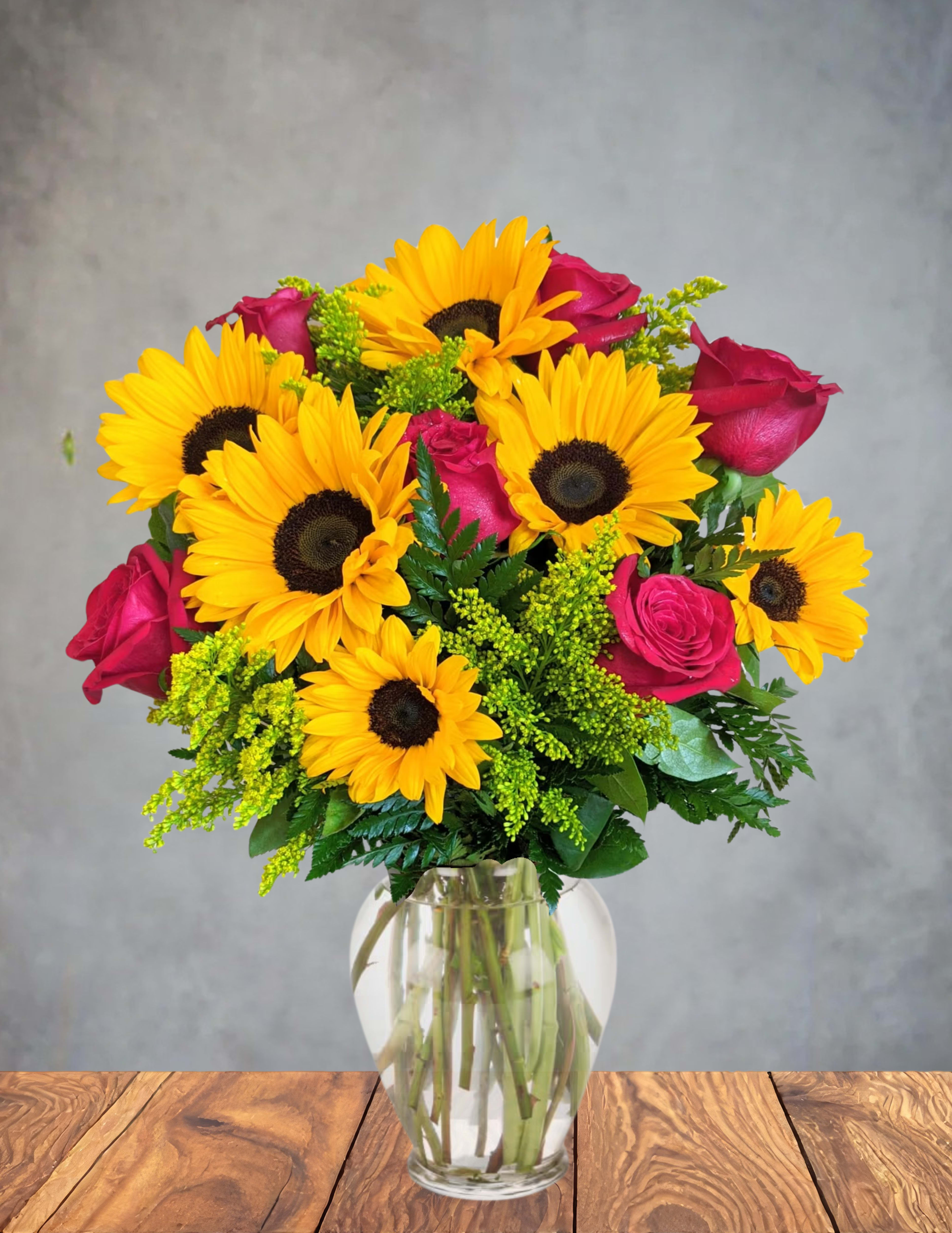 Sippin sunsets - A warm, easy‑going arrangement that glows like the last light of day. Golden sunflowers rise like little suns, their bright faces radiating comfort and joy. Deep red roses swirl through the bouquet like the rich hues of a sangria‑colored sky, adding a slow‑burn romance. Soft greenery tucks everything together, giving it that relaxed, front‑porch, end‑of‑day charm.  The whole piece feels like a toast to simple pleasures—warm evenings, good company, and the kind of sunsets you want to sip slowly.