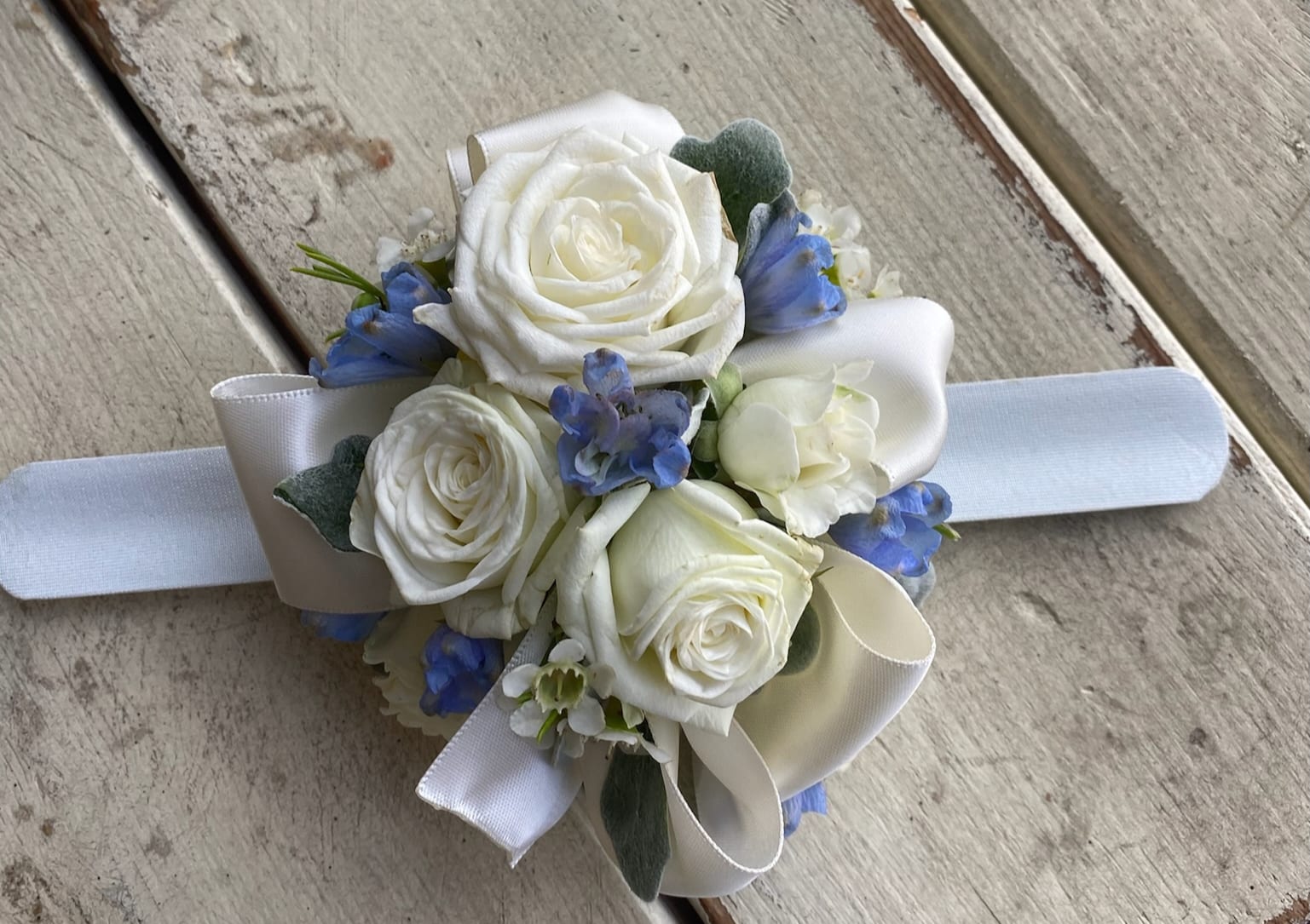 Wrist Corsage #2- Roses and Delphinium - White spray roses, light blue delphinium, filler flowers and greenery on a wrist band. Please specify ribbon color