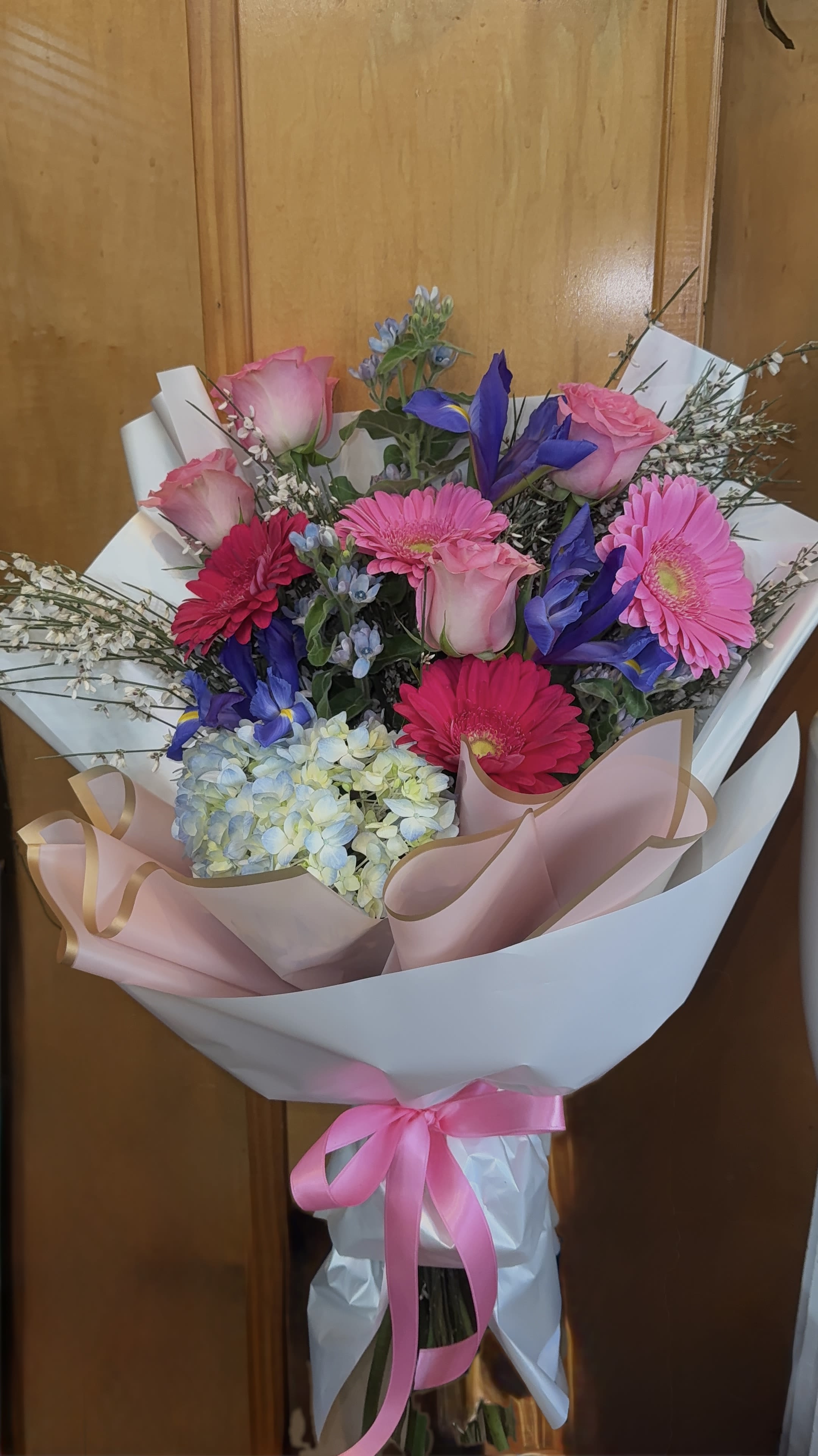 Spring kiss  - A lively and elegant hand-tied bouquet bursting with shades of pink, blush, soft blue, and violet. Featuring cheerful gerberas, romantic roses, delicate hydrangea, and graceful irises, this arrangement is wrapped in soft blush and white paper and finished with a satin pink ribbon. Fresh, feminine, and full of charm, it’s perfect for birthdays, congratulations, anniversaries, or simply to brighten someone’s day. Please note: This bouquet is sold without a vase. 