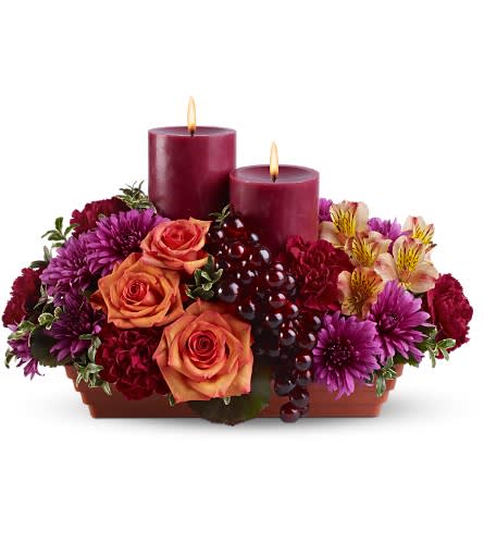 Bordeaux by Candlelight - There's nothing more wonderful than sipping a glass of red wine by candlelight; and this glamorous display is that lovely moment brought to life with flowers! Two pillar candles and glossy grapes complete the picture. An exceptional holiday centerpiece.    A mix of fresh flowers such as roses, carnations and cushion spray chrysanthemums - in shades of orange, maroon, lavender and peach â is accented with faux grapes and arranged in a tray with two burgundy pillar candles.    Approximately 9" (W) x 13" (H)    Orientation: All-Around        As Shown : TFWEB457    