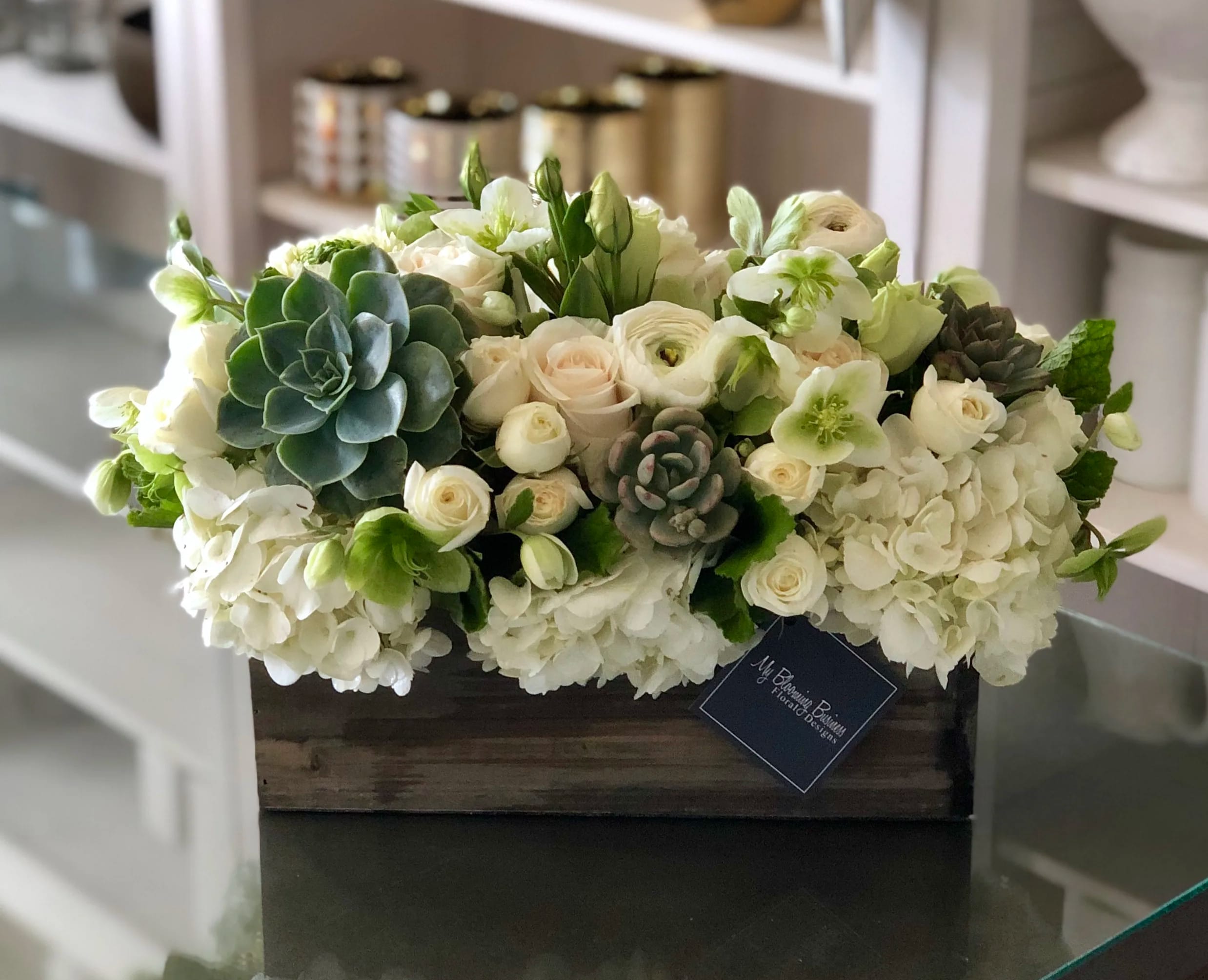 The Mulholland  - The Mulholland is your perfect blend of rustic and elegance! This beautiful and unique box arrangement flaunts white flowers, lisianthus, spray roses, and other seasonal florals, with some succulents to tie it all together. Named after the Winding Roads of Mulholland Drive, this arrangement offers a touch of glamour and evening sky vibes. Perfect for your friends or family, this arrangement brings sophisticated and intriguing floral vibes to any room. Give your loved ones something truly special with the Mulholland!