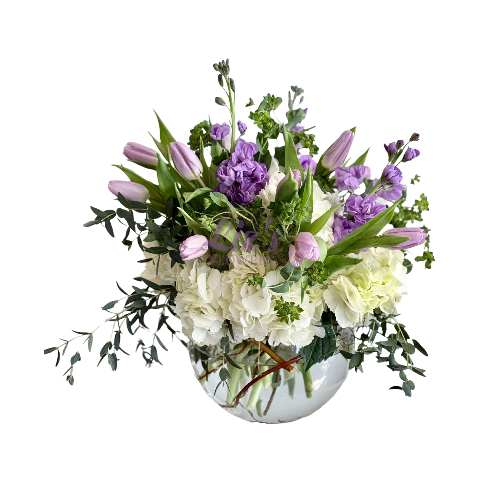 Lavender Whisper Bouquet - A fresh mix of purple tulips and stocks, soft white hydrangeas, and leafy greens arranged in a clear round vase.