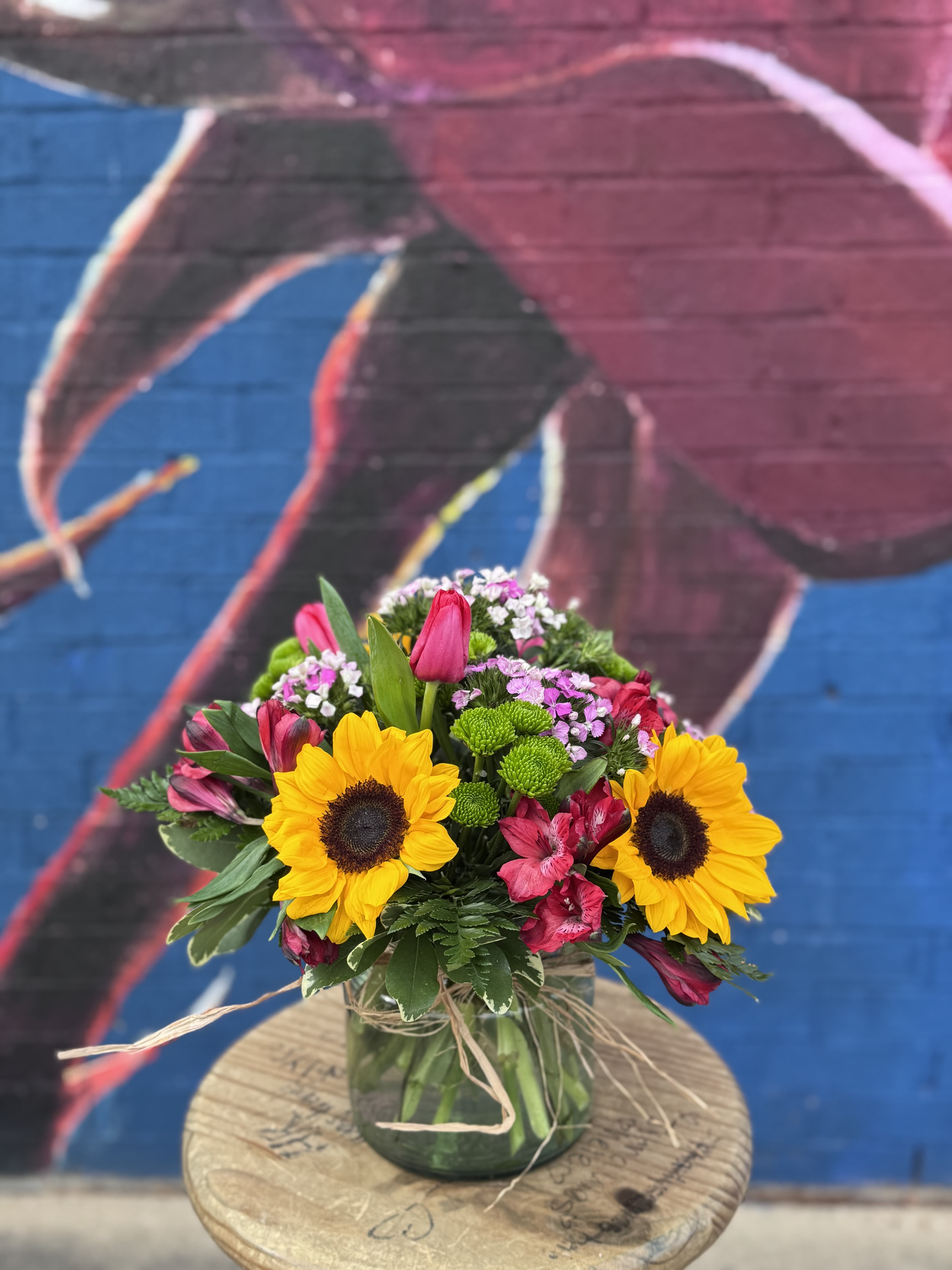 Sunshine of My Life - Bright, joyful, and full of heart—Sunshine of My Life is a Valentine’s arrangement that celebrates the person who lights up your world. Featuring cheerful sunflowers paired with vibrant tulips, playful button mums, soft alstroemeria, and pops of statice, this design is bursting with color and warmth.  Arranged in a clear glass vase accented with natural raffia, Sunshine of My Life is perfect for Valentine’s Day when love feels happy, easy, and full of joy.