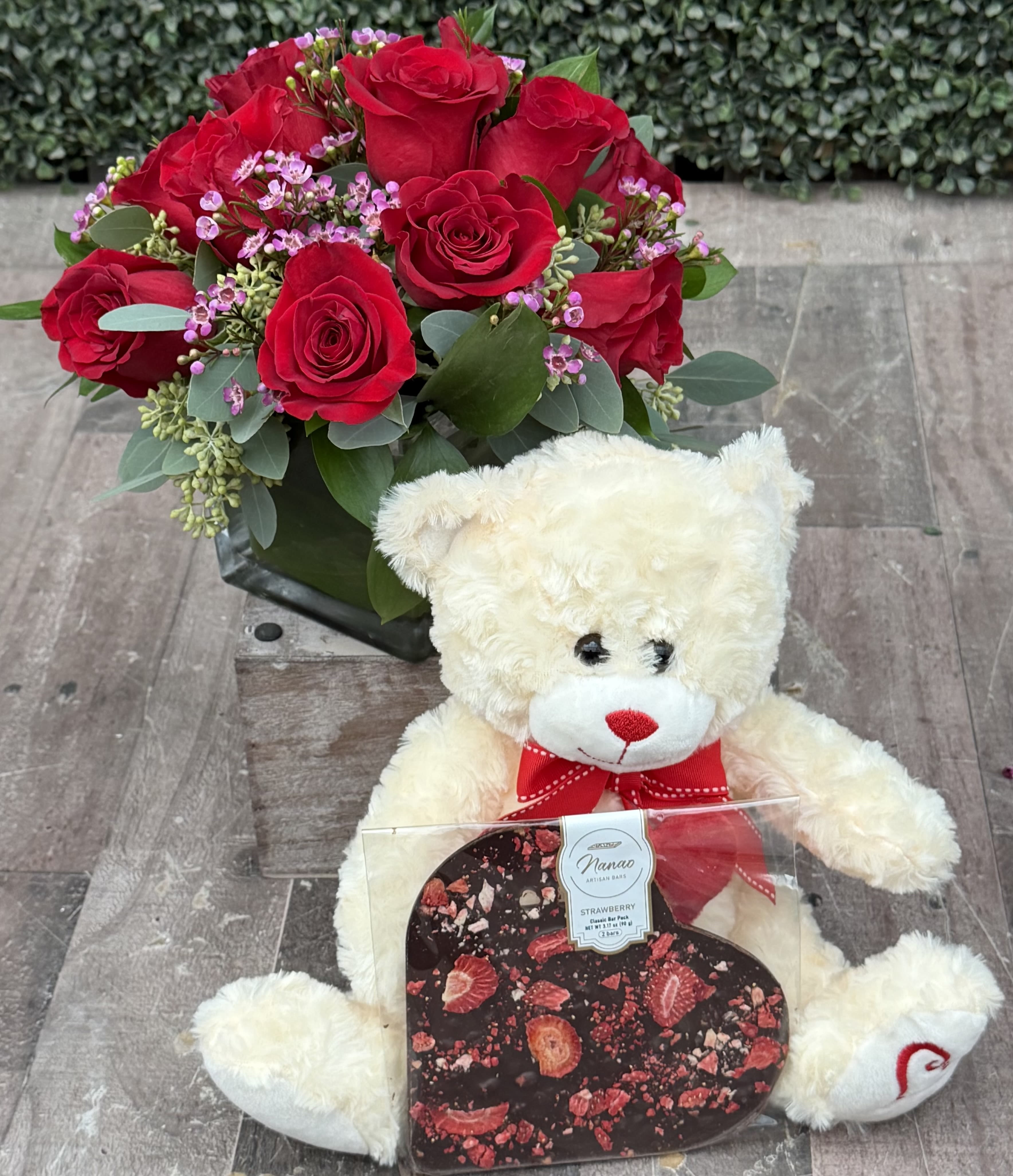 CLUSTER OF LOVE (RED) VALENTINE'S GIFT SET - This clustered arrangement comes designed with roses, seeded eucalyptus, wax flower and assorted greenery in a cube wrapped with a leaf.  It is paired with a Valentine's Teddy bear and a heart shaped artisan chocolate. Bear and chocolate come in a decorative window bag. 