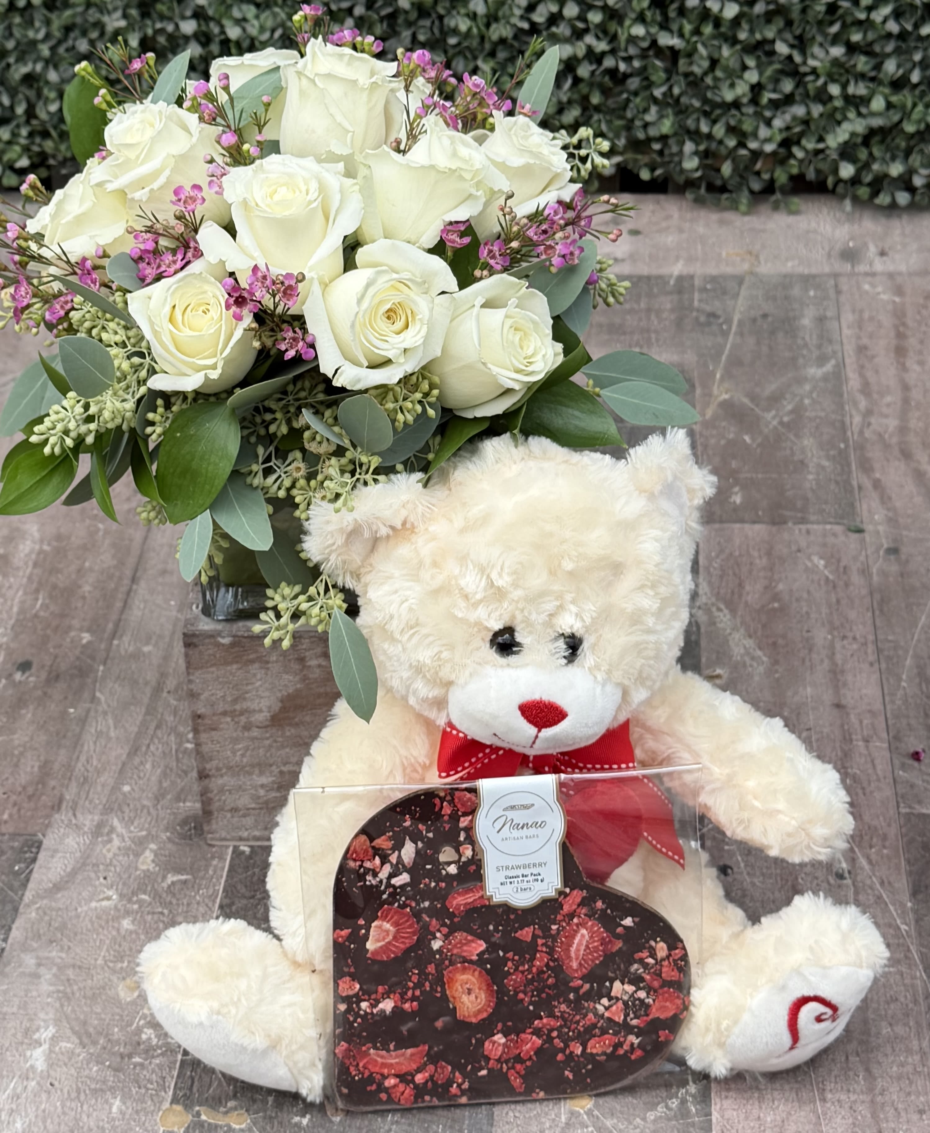 CLUSTER OF LOVE (WHITE) VALENTINE'S GIFT SET - This clustered arrangement comes designed with roses, seeded eucalyptus, wax flower and assorted greenery in a cube wrapped with a leaf.  It is paired with a Valentine's Teddy bear and a heart shaped artisan chocolate. Bear and chocolate come in a decorative window bag. 
