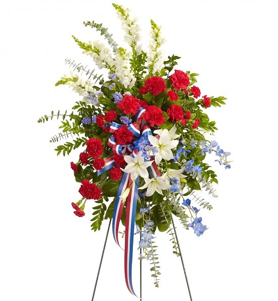 Sacred Duty Standing Spray - Adorned with yards of multicolored patriotic ribbon, this easel-mounted standing spray features flowers such as red carnations, white asiatic lilies and snapdragons, and blue delphinium, along with an assortment of greens.