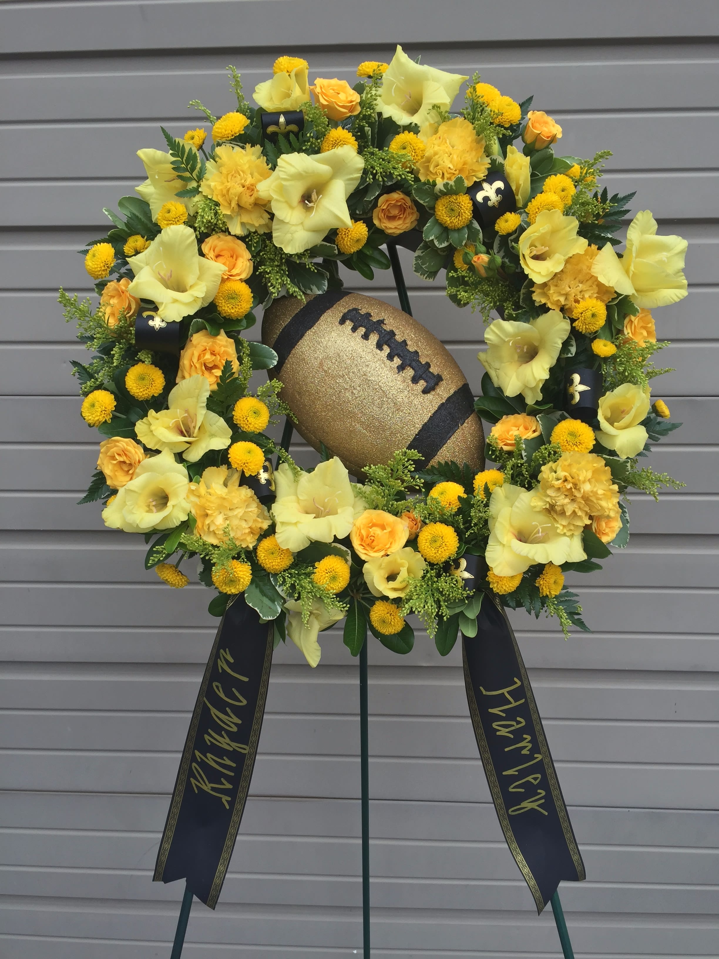 Saints Fan Remembrance Wreath - Celebrate your loved one's favorite team with this Saint's themed open circle standing easel. Made with all yellow gladiolus, spray roses, cushion poms, and carnations and accented with Saints ribbon, a golden glitter football, and two banners with a message of your choice. Please include in your order what message you would like printed on each of the banners.