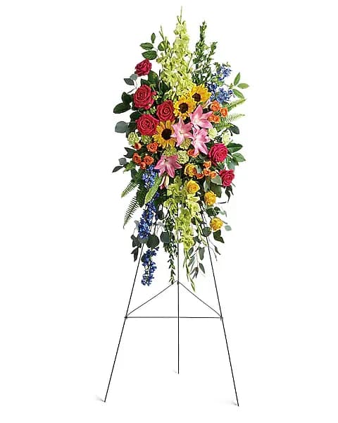 Love Lives On Spray - Bursting with joy, this radiant spray of rainbow blooms is an exuberant tribute to never-ending love.  This spray features hot pink roses, yellow roses, orange spray roses, pink asiatic lilies, green gladioli, green carnations, medium yellow sunflowers, blue delphinium, bupleurum, myrtle, sword fern, silver dollar eucalyptus, and lemon leaf. Delivered on a wire easel.
