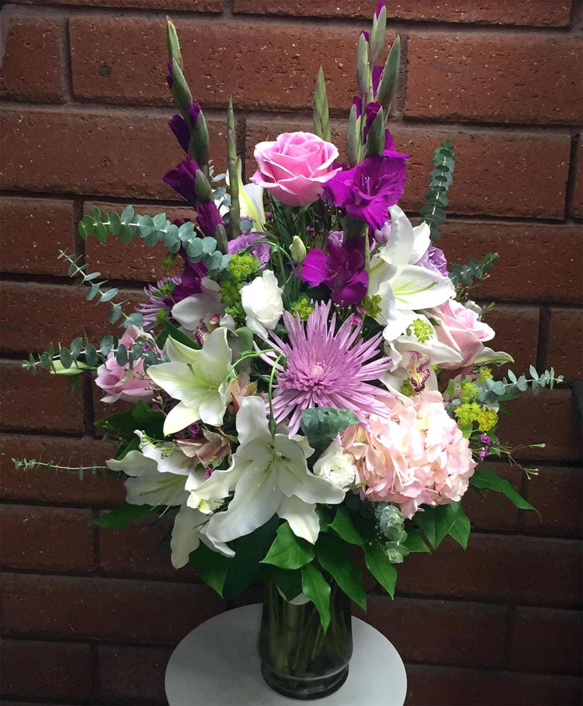 Amethyst Moon - Intriguing hues of pink, white, lavender, and purple come together to create an atmosphere of calm and quiet elegance. The Amethyst Moon arrangement features a luxurious mix of roses, fragrant Crystal Blanca lilies, striking gladiolus, cheerful chrysanthemums, lush hydrangea, alstroemeria, and exotic cymbidium orchids. Thoughtfully designed to soothe the senses, this bouquet offers a peaceful escape from the rhythm of everyday life.  Standard size measures approximately 18" (W) x 29" (H). Deluxe and Premium versions are larger, featuring additional premium blooms and upgraded clear glass vases for a fuller, more dramatic presentation.  Available Sizes:  • Standard – One-sided presentation style arrangement in a clear glass vase  • Deluxe – Full, round style design with additional blooms in a glass vase  • Premium – Jumbo full, round style design with the most abundant floral display in a larger glass vase  Please Note: Upon delivery, most lilies will arrive in bud form to ensure maximum freshness and longevity. As lilies open, gently remove the stamens to enhance flower life and prevent pollen staining.  Care Tips: Check the water level daily and keep the vase filled with fresh, clean water. Replace the water every 2–3 days and trim the stems with a sharp cut to enjoy long-lasting beauty.  Elegant, soothing, and timeless, Amethyst Moon is a perfect choice for moments that call for peace, comfort, and refined beauty.