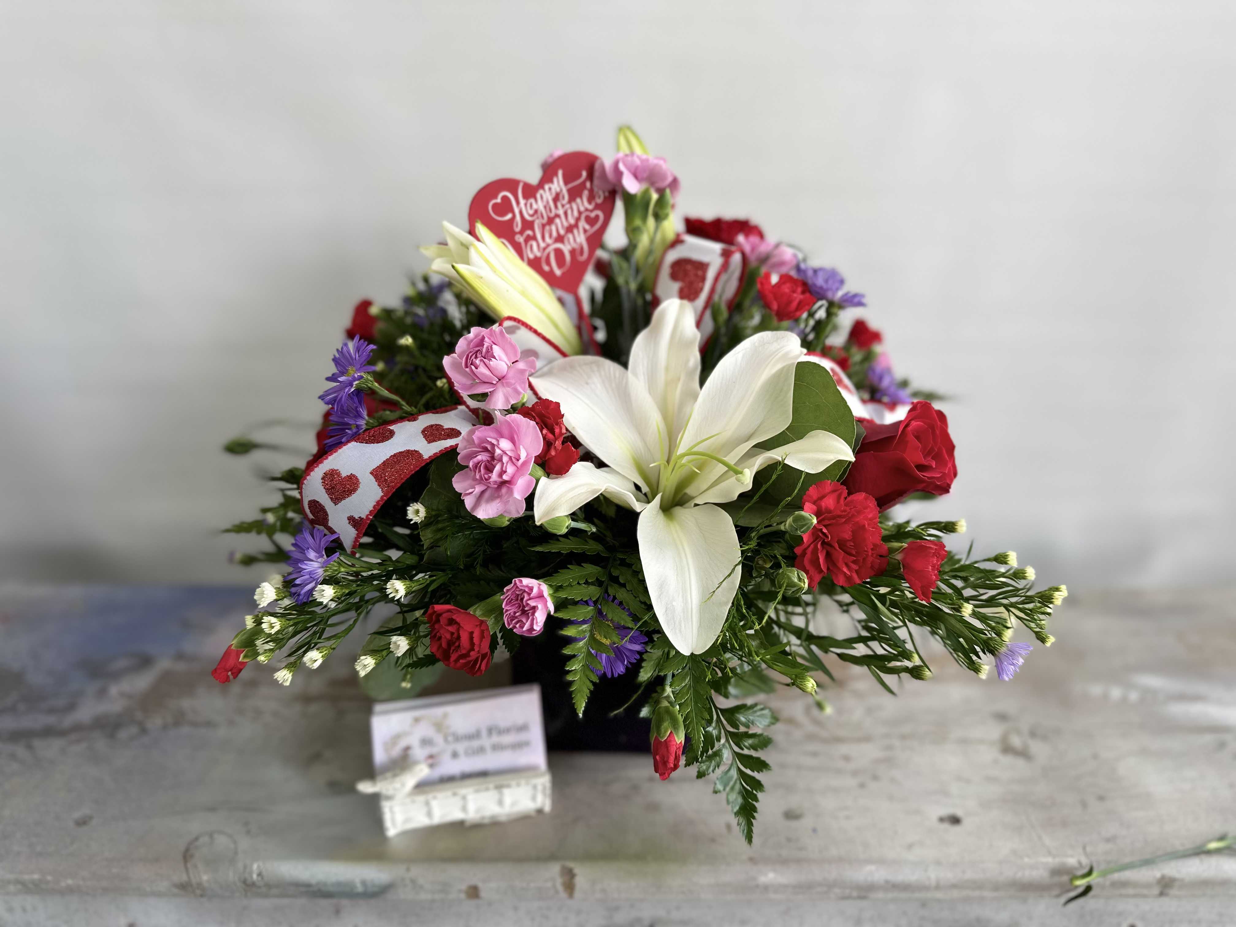 Forever Yours - A stunning mix of roses, lilies, and vibrant flowers in Valentine's Day colors. Arrangement comes in Golden Hearts cylinder. While supplies last. Flowers and containers are subject to substitution.