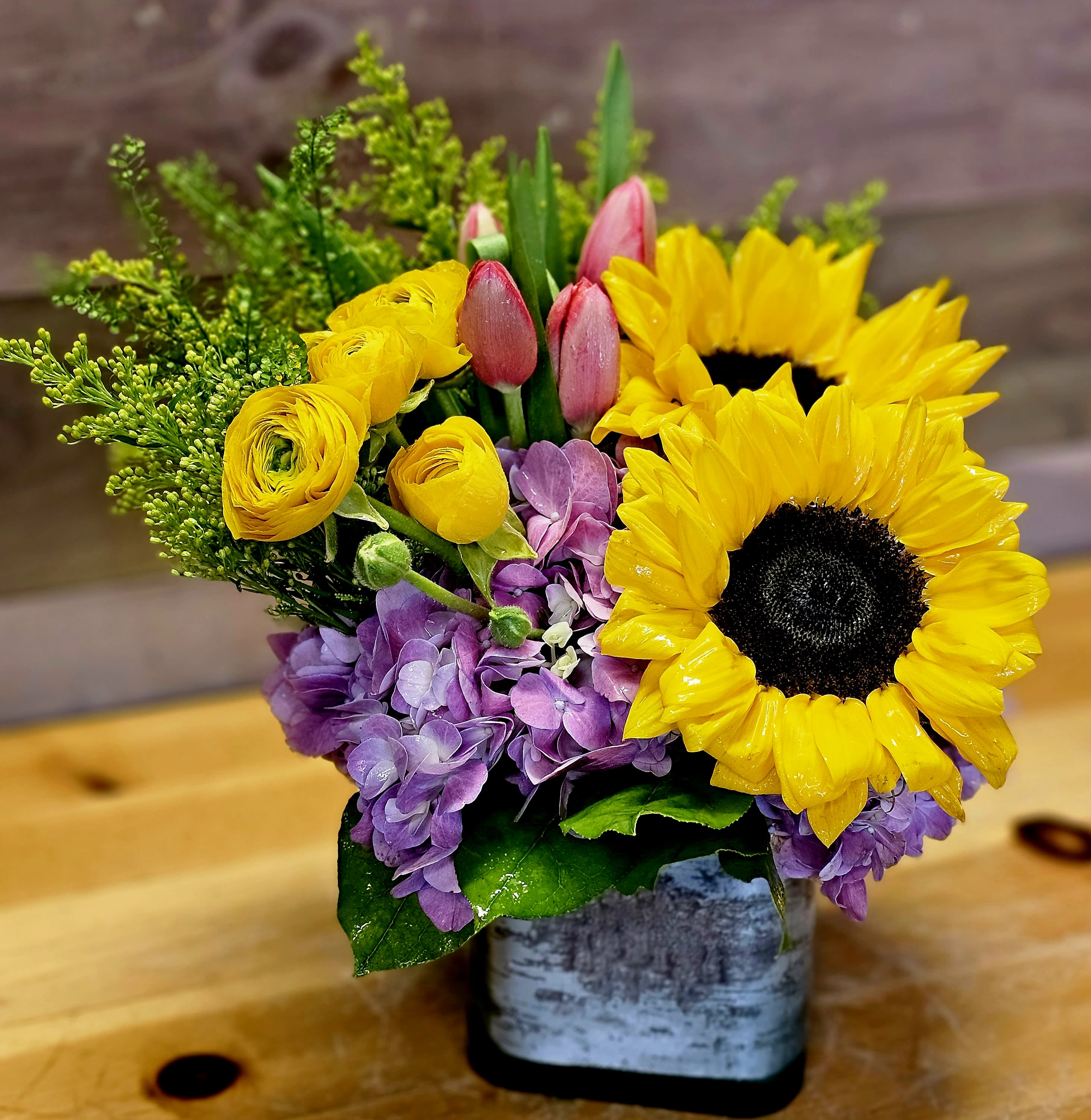 Cuteness  - Send this to the cutie in your life! Great for a birthday, or just cheering somebody up! This summertime arrangement with sunflowers, tulips, and spray roses lights up any space. Color of flowers will change, for what is stock. Container can and will vary upon stock. Flowers can change with season and current stock. 