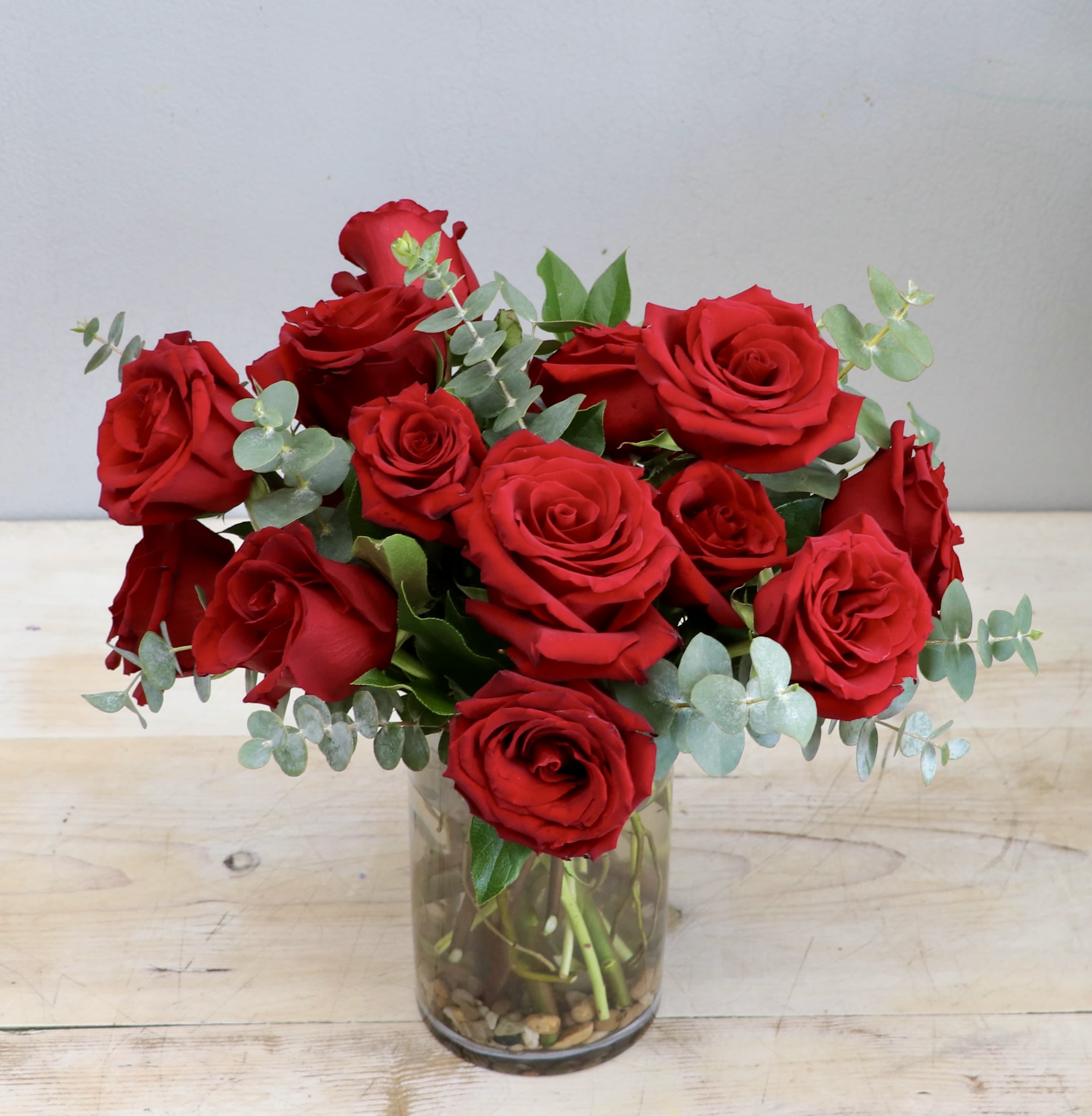 Valentine Red - WEHO - This classic arrangement features a lush gathering of deep red roses nestled among delicate sprigs green foliage. Presented in a sophisticated glass vase, it serves as a timeless symbol of passionate love and elegance for Valentine's Day.  