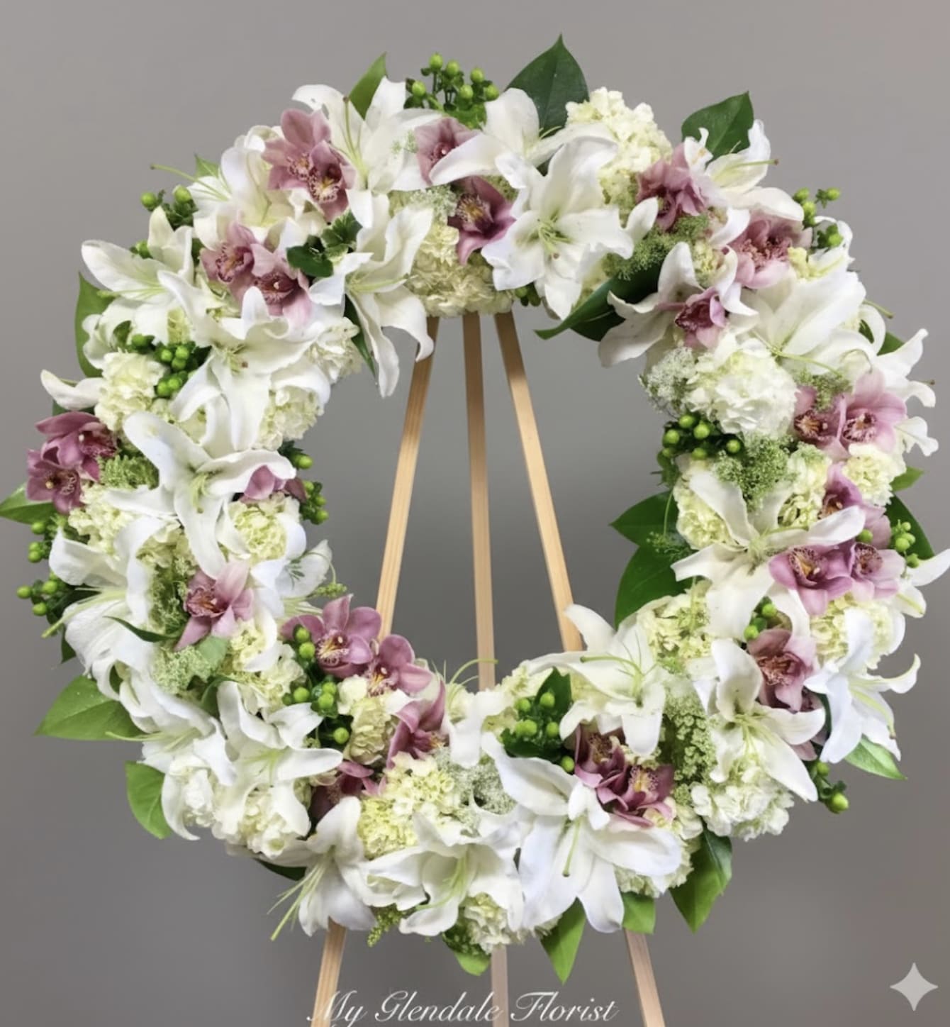 Orchid and Lily Wreath  - Orchids, lilies, and hydrangeas wreath. Standard size measures approximately 30 inches wide. Deluxe measure approximately 36 inches wide. Premium measures approximately 42 inches wide.    For best fit, we recommend a message of 40 characters or less for banners.     We include easel, printed banner, and delivery (some fees may apply). An additional banner can be added for $15  