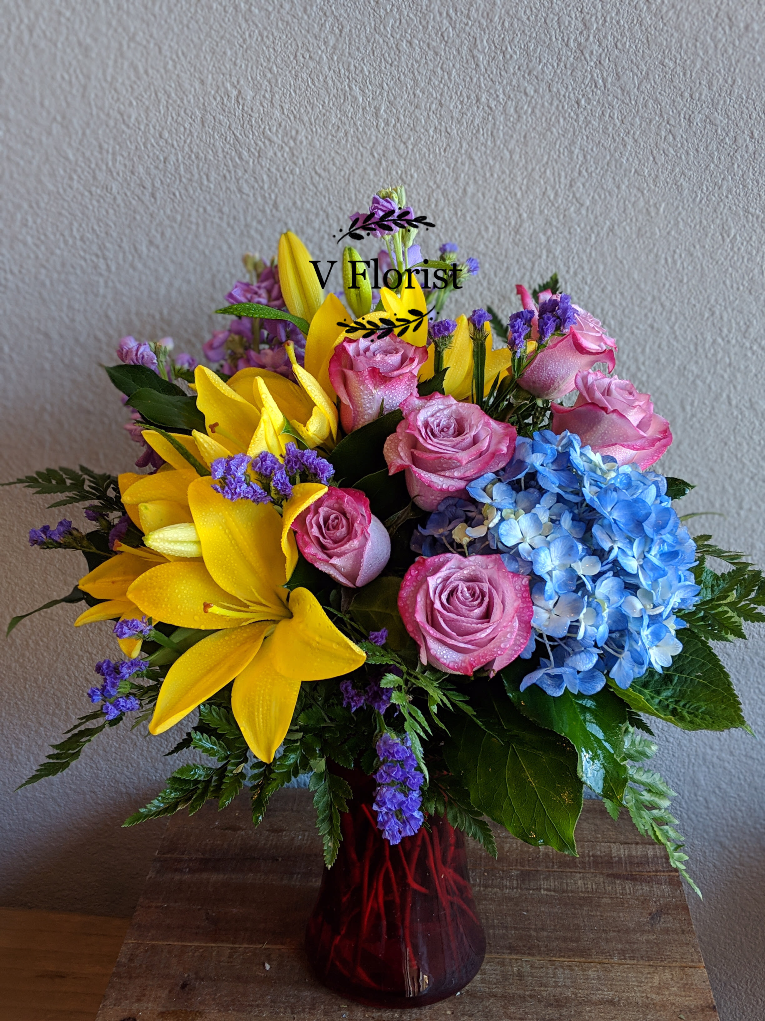 Spring Equinox Bouquet - Designer's choice of blue hydrangea, yellow lilies, lavender roses, statice , and lavender stocks in a red vase.