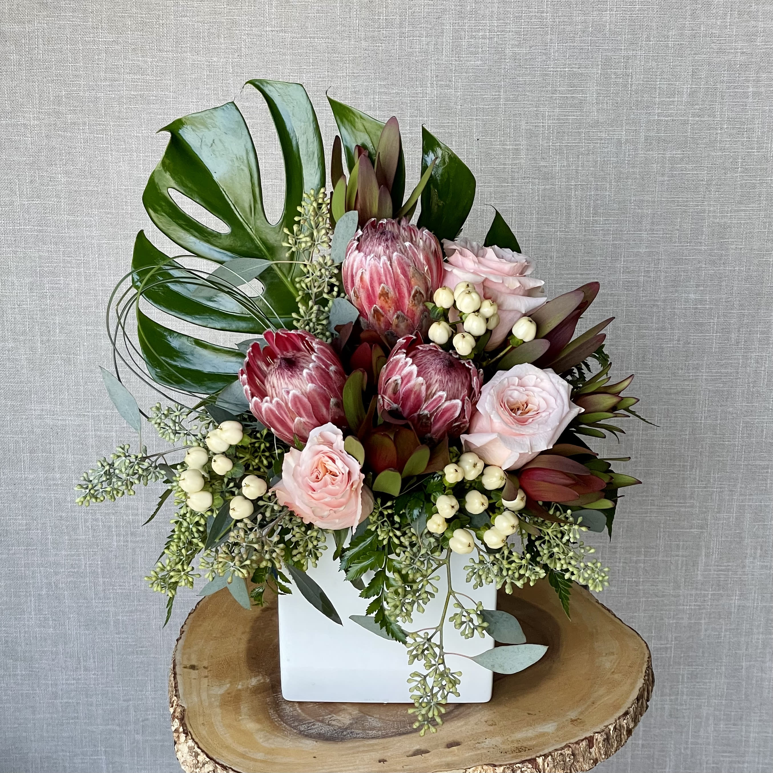 Tropical Blush Harmony (SF402) - This floral arrangement features Shimmer roses, proteas, coffeeberries, and safari sunset, seeded eucalyptus, complemented by a monstera leaf, all elegantly displayed in a contemporary white ceramic square vase. 