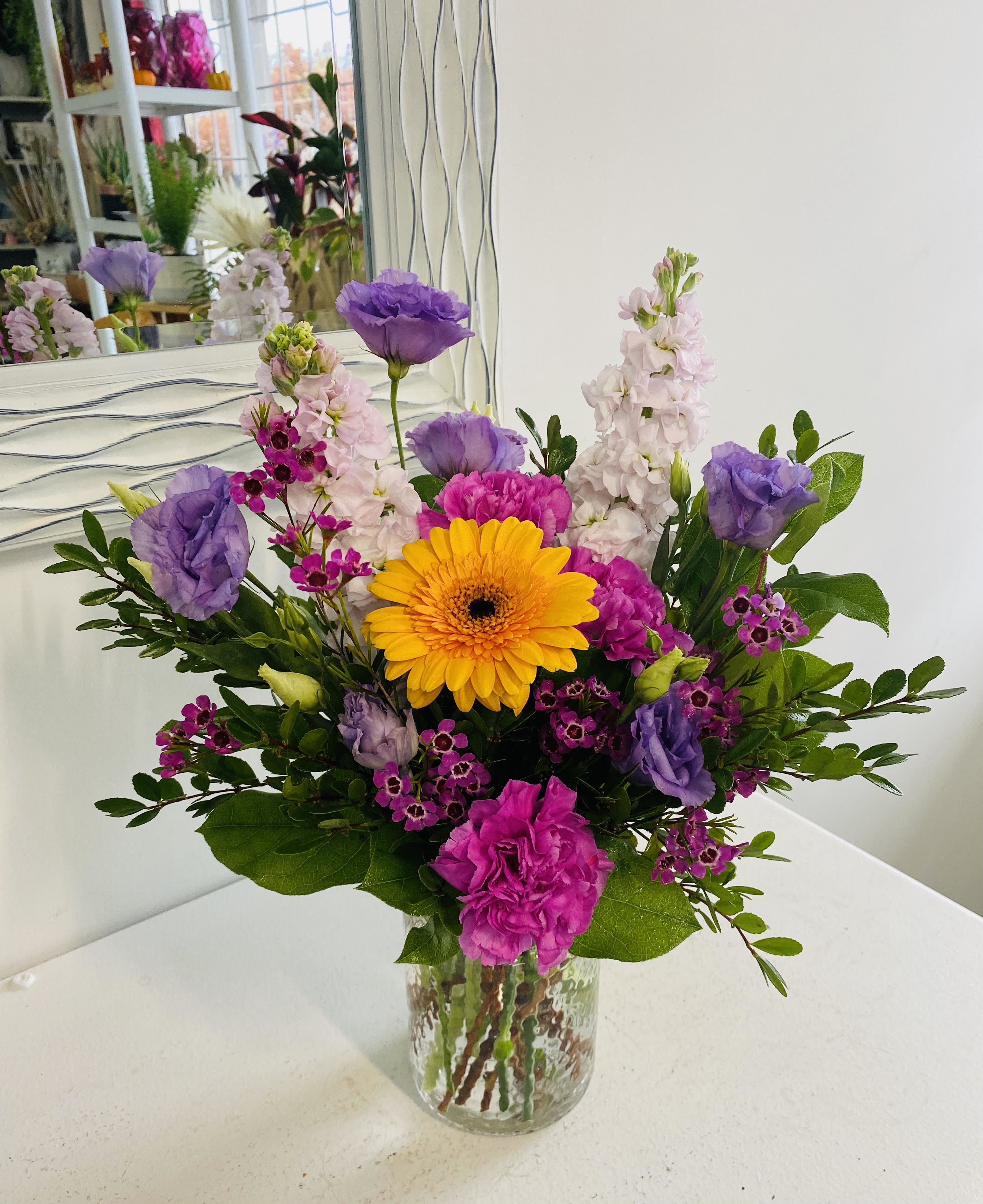 Heaven Scent - This cheerful bouquet features a sunny yellow gerbera daisy at its center, surrounded by soft lavender lisianthus, bright pink carnations, delicate white stock, and vibrant magenta waxflowers. Set in a clear textured glass vase with lush greenery, it’s a fresh and uplifting display perfect for brightening any room.