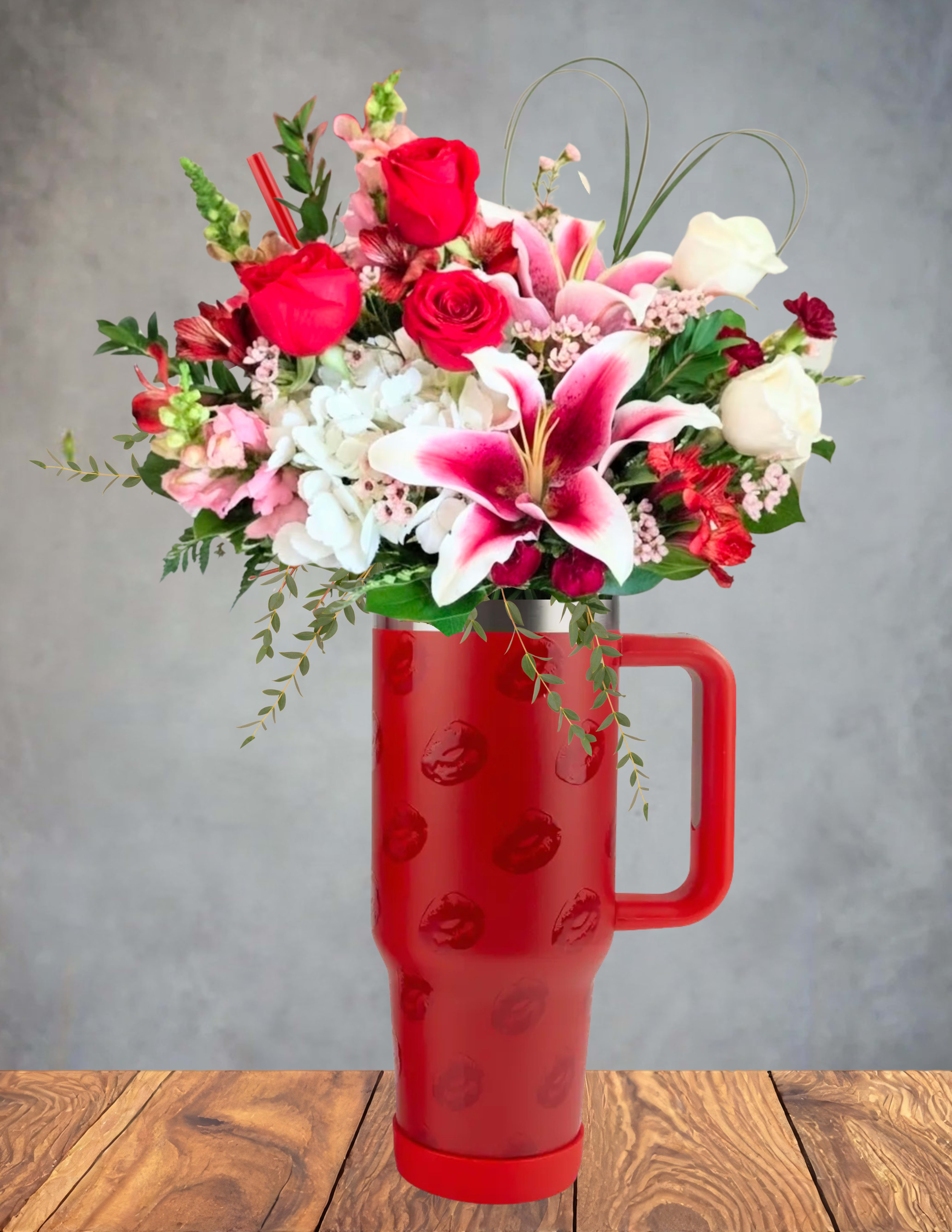 Cupids first kiss  - A playful spark of romance captured in bloom. This charming arrangement bursts from a red lip‑embossed travel mug, as if Cupid himself left a kiss behind. Red and white roses mingle with soft pink lilies, hydrangea, and flirty seasonal blooms, creating a lush swirl of color and texture.  Perfect for secret admirers, new beginnings, or anyone who deserves a little love‑struck magic. And when the flowers fade, the keepsake mug keeps the moment alive with every sip.