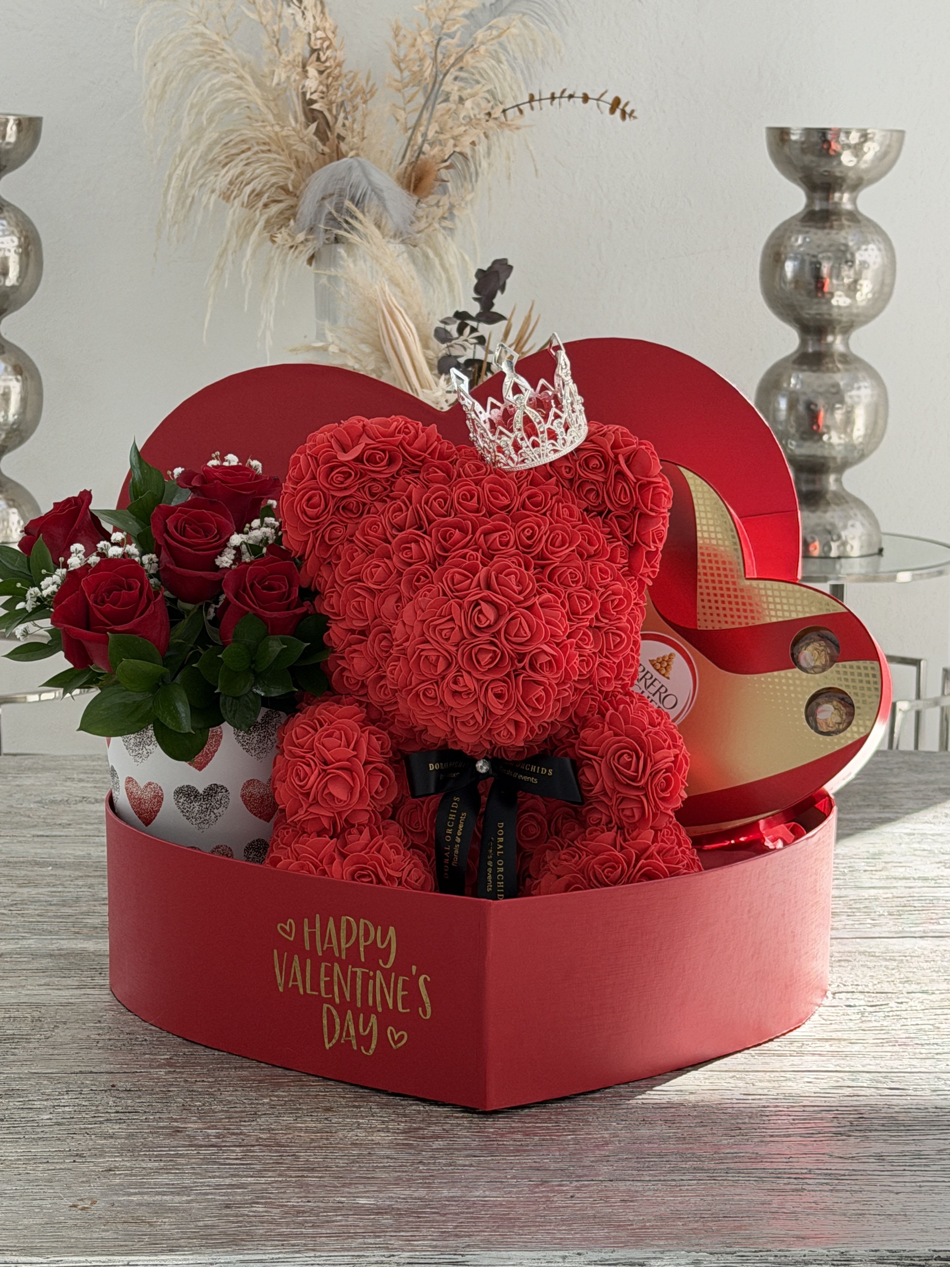 Royal Rose Bear Luxe Box -  Please add the recipient’s name in the Special Instructions at checkout for personalization.   Make a bold statement of love with our Royal Rose Bear Luxe Box, a show-stopping Valentine’s keepsake designed to impress. This handcrafted red rose bear is crowned with a sparkling silver tiara, symbolizing love, devotion, and royalty. Nestled inside a customizable heart-shaped box, this arrangement includes fresh premium red roses and an elegant Ferrero Rocher chocolate heart for the ultimate romantic touch.  The box itself can be personalized to make your gift truly one-of-a-kind, turning this piece into a lasting memory long after Valentine’s Day. Perfect for anniversaries, proposals, or unforgettable Valentine surprises.