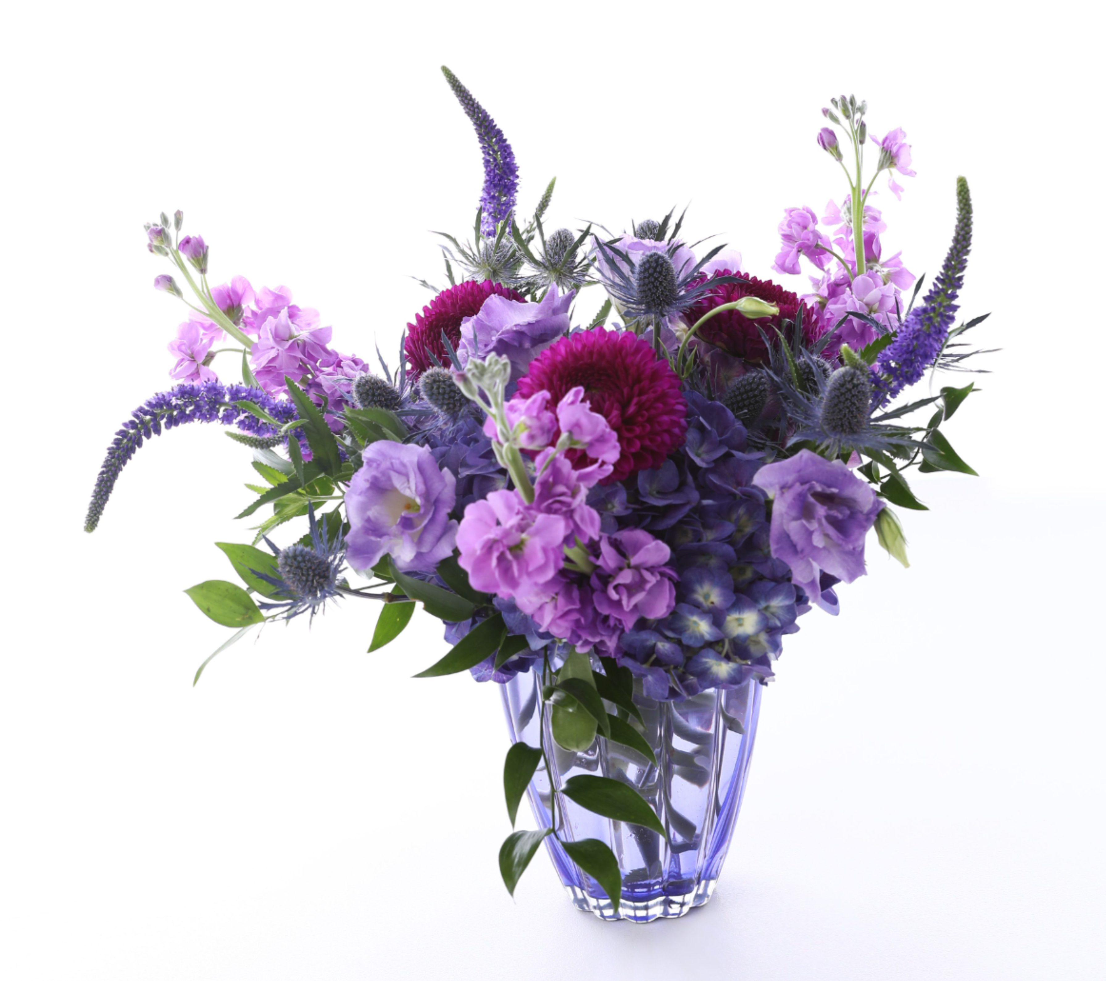 LAVENDAR ROMANCE - WE TOOK ALL THE SHADES OF PURPLE FLOWERS AND COMBINED THEM IN THIS PURPLE GLASS VASE. LAVENDER STOCK AND LISIANTHUS STAND OUT AMONGST THE STRONGER PURPLE HUES OF HYDRANGEA, VERONICA, AND DAHLIAS. WE THEN ADDED BLUE THISTLE TO FINISH IT OFF. 