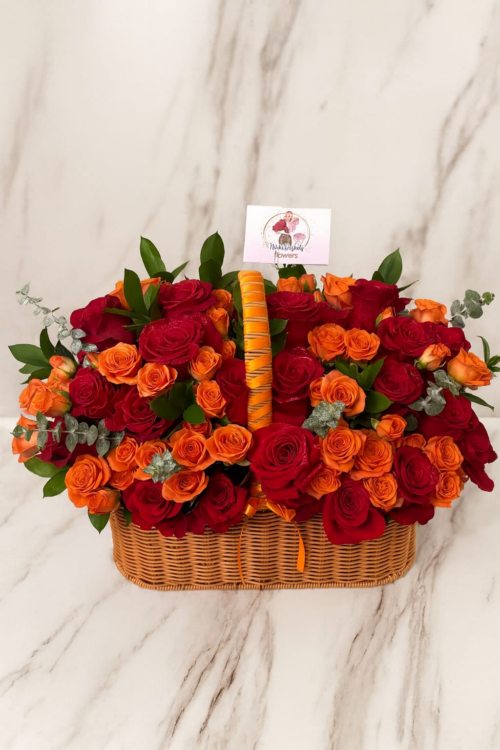 Luxury Rose Basket Orange - Celebrate in style with this stunning handcrafted basket overflowing with vibrant red roses and elegant orange spray roses, beautifully accented with eucalyptus and lush greenery. Presented in a classic woven basket, this arrangement is perfect for birthdays, anniversaries, or any special occasion. A timeless gift that radiates elegance and charm.