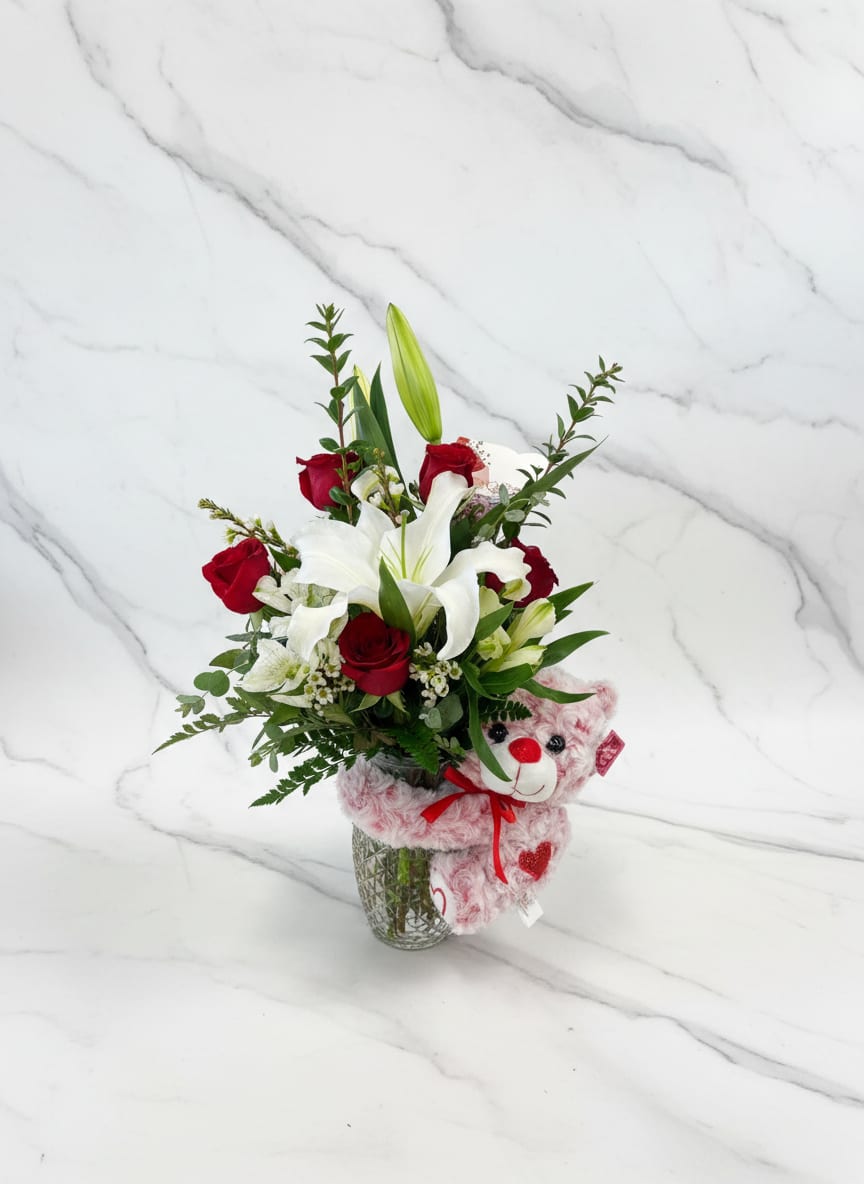 Love you Beary Much - A charming vase arrangement with a bear hugging the vase, designed with red roses, white oriental lilies, white alstroemeria, wax flower, and lush greenery. A perfect mix of romance and sweetness.