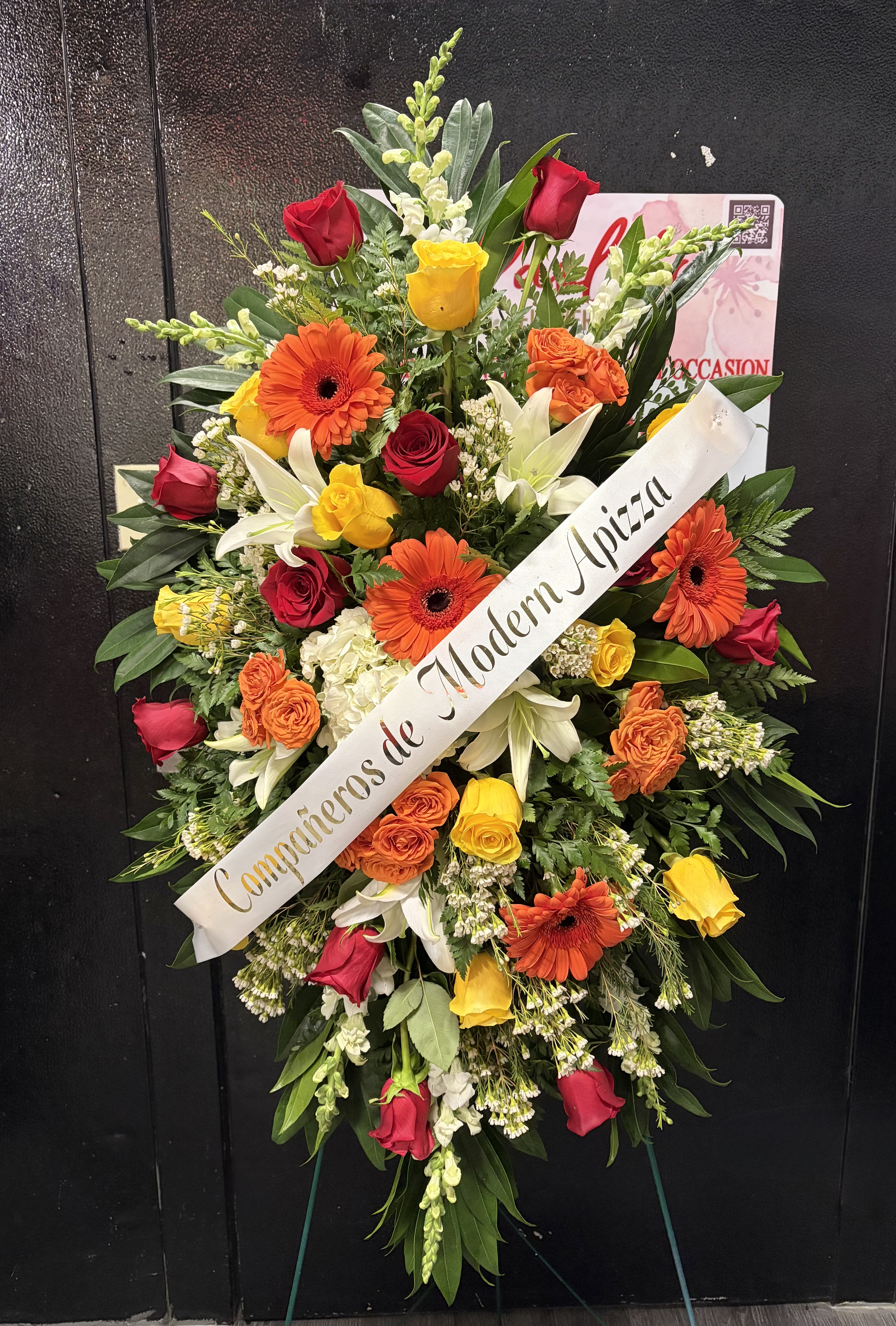 Vibrant Memories Standing Spray - Celebrate a life full of color, warmth, and cherished memories with our Vibrant Memories Standing Spray. This bold and uplifting arrangement features a radiant blend of red and yellow roses, orange gerbera daisies, white lilies, hydrangea accents, and lush greenery—symbolizing love, gratitude, and joyful remembrance.  Designed to bring comfort and light during times of loss, this standing spray is thoughtfully handcrafted by our florists and displayed on an easel, making it a meaningful tribute for funeral services, memorials, or celebrations of life. Ribbon message customization is available.