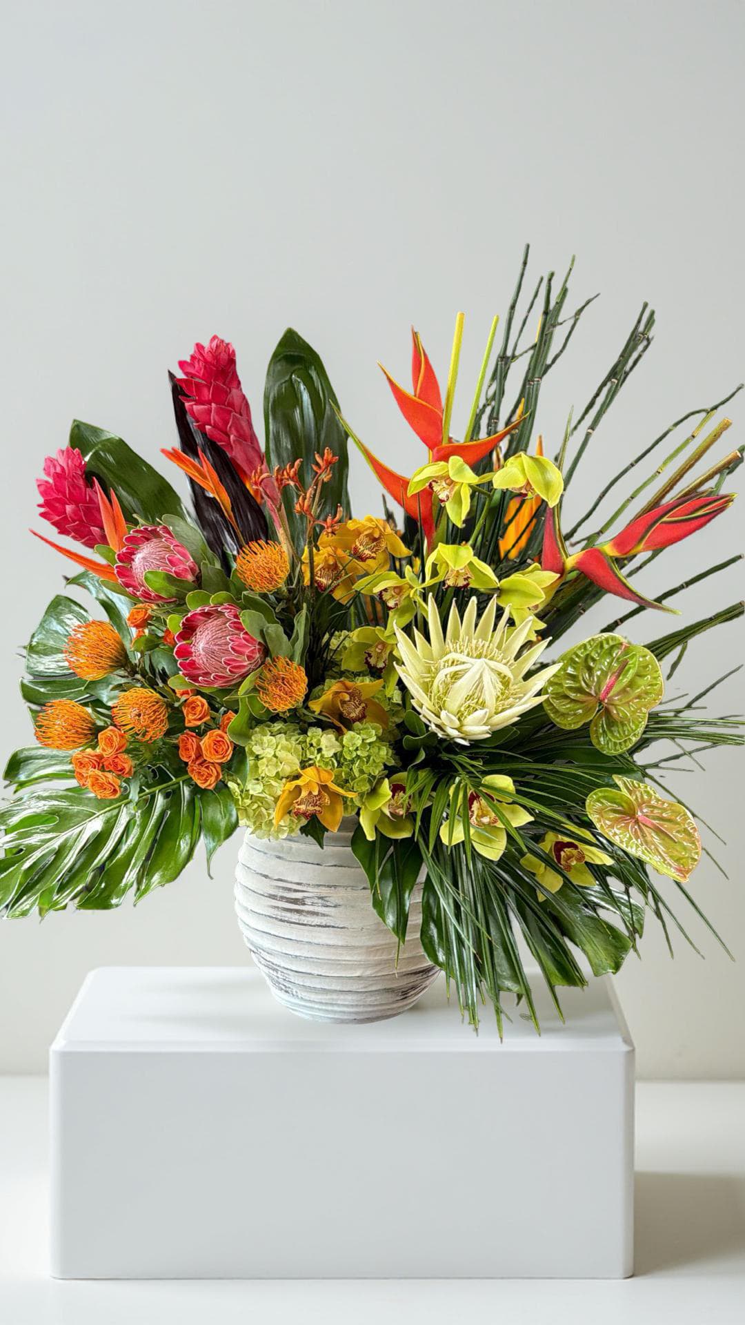 Tropical Like It's Hot - Immerse yourself in the extraordinary beauty of the tropics. This captivating, large arrangement brings the drama of the islands to life, combining the vibrant colors and unique shapes of exotic blooms like protea, heliconia, anthuriums, and orchids.