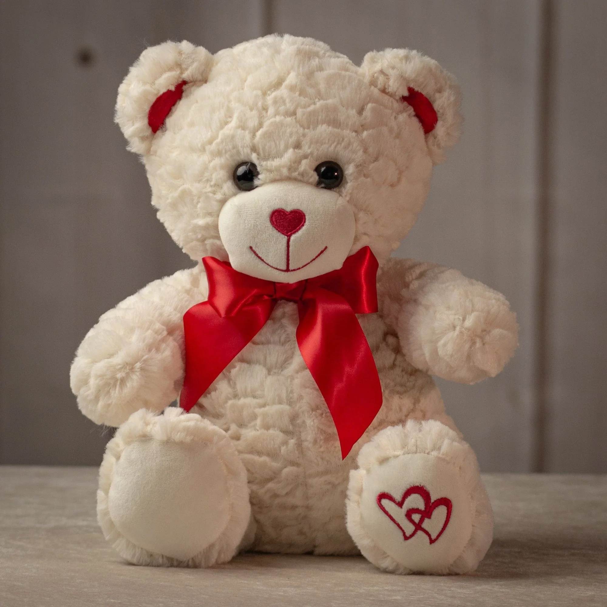 12" Heart Print Valentine Bear - This ivory-colored teddy bear exudes sweetness with its heart-shaped red nose, matching red satin bow, and an embroidered heart design on its paw. Its soft, textured fur makes it a charming and huggable gift for Valentine's Day, spreading love with every cuddle. Size - 12" inches tall while sitting from head to bottom Feel - Soft and cuddly, stuffed with hypoallergenic fiber fill