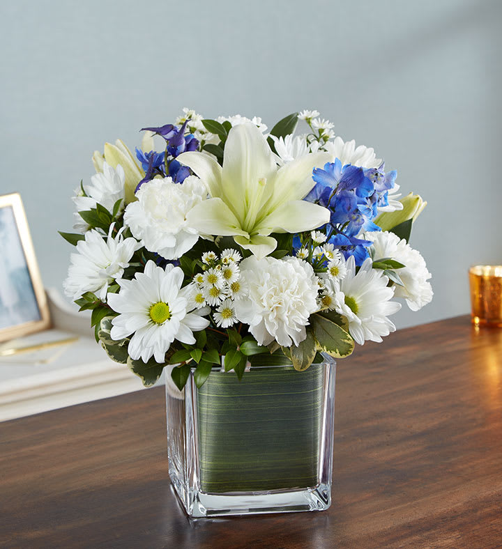 Healing Tears Blue &amp; White - Soothe their tears as they mourn the loss of a loved one with the serenity of blue and white. Our sympathy arrangement of fresh blue delphinium, white roses and lilies, expertly gathered together in a clear glass cube lined with Ti leaf ribbon, makes for an exquisite gesture of comfort and healing.