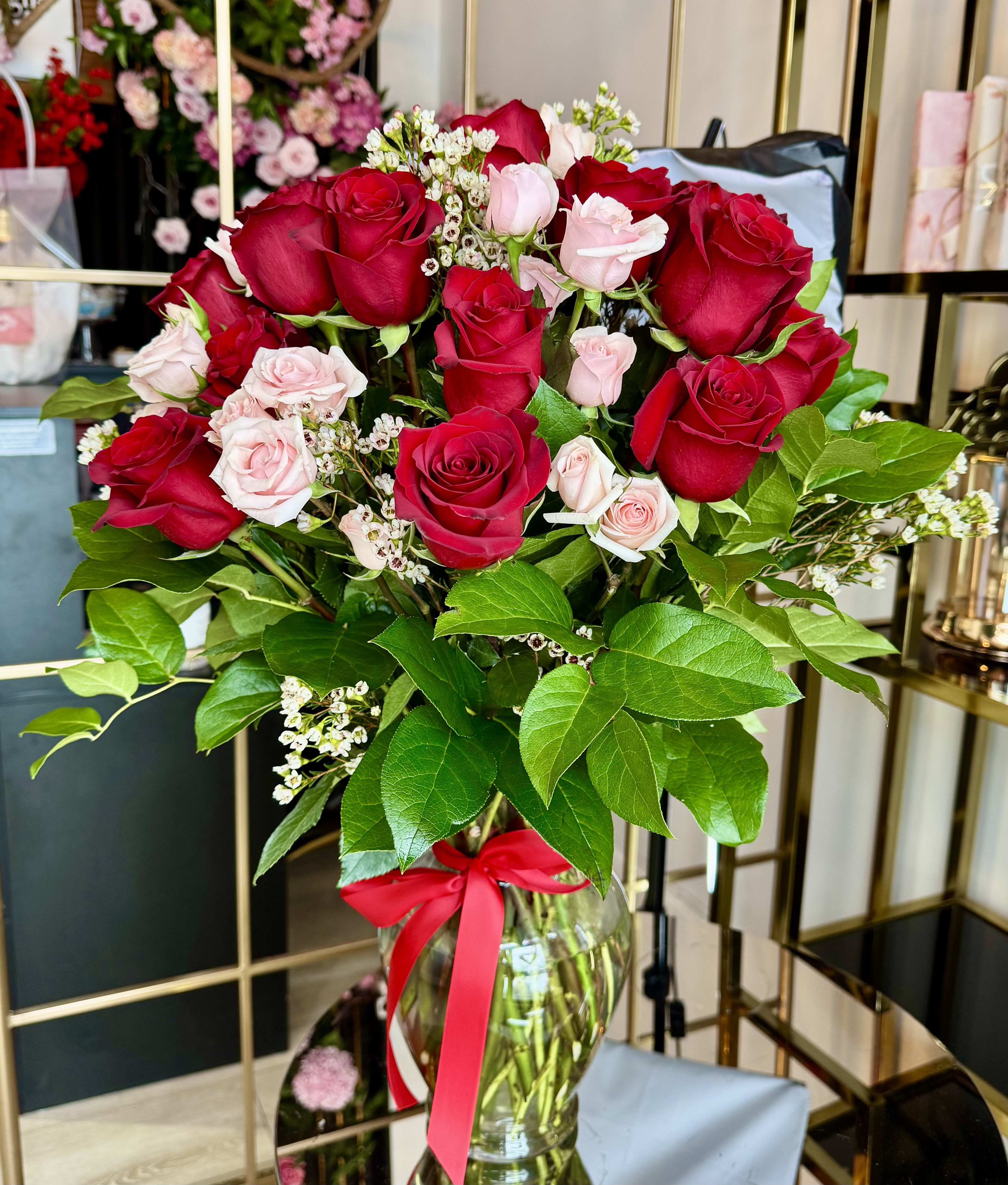 Crimson Blush Rose Vase Arrangement - timeless romantic design featuring 12 premium red roses paired with delicate pink spray roses, lush seasonal greens, and soft white accents. Designed in a clear glass vase and finished with a red ribbon, Crimson Blush is the perfect expression of love for Valentine’s Day, anniversaries, or any romantic occasion.