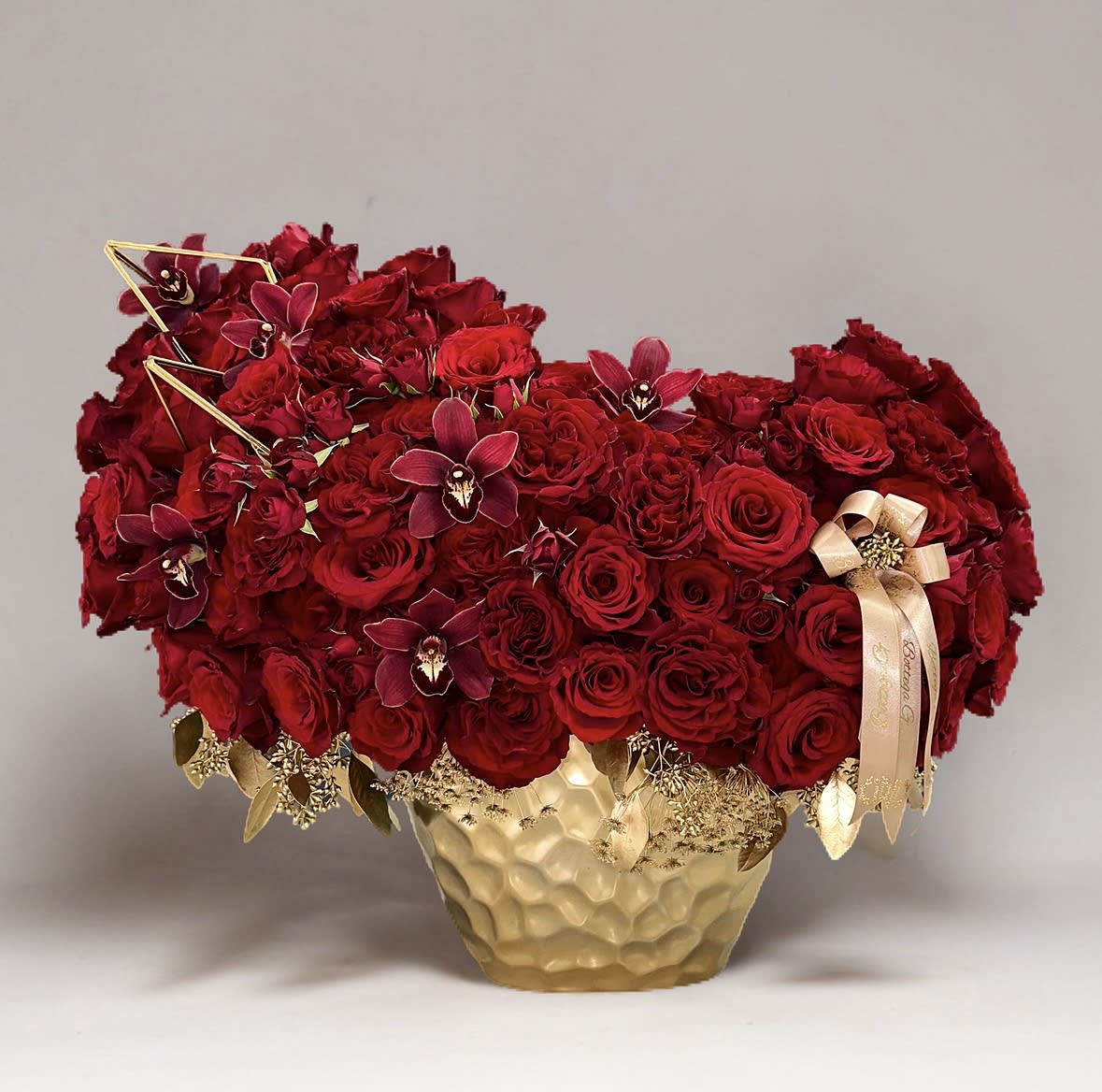 Luxe Red Rose Ensemble - The arrangement exudes sophistication with a lush collection of vibrant, velvety red roses and beautiful moth orchids. Their rich color symbolizes love, passion, and elegance.  Now, imagine these gorgeous red roses nestled within a sleek, opulent gold vase. The vase itself adds a regal and luxurious element to the arrangement. Its lustrous golden surface complements the vivid red of the roses, creating a striking contrast that captivates the eye.  The arrangement stands tall and proud, the roses delicately arranged to create a sense of natural flow and effortless beauty. The gold vase adds a touch of glamour and sophistication, elevating the entire arrangement to a level of refined elegance.  Placed as a centerpiece or displayed in a prominent area, this arrangement of red roses in a gold vase becomes a focal point, exuding warmth, romance, and timeless charm.