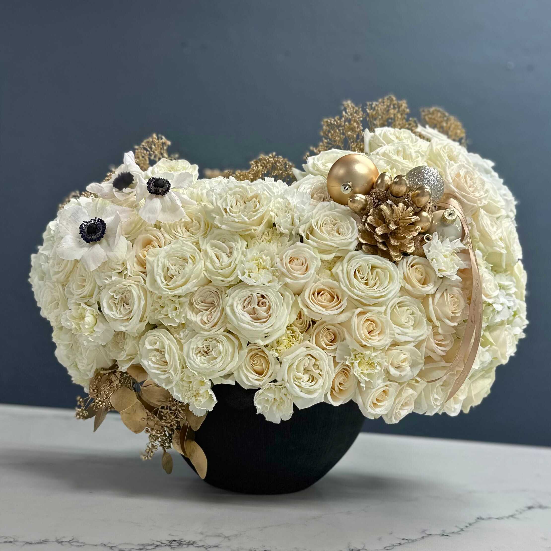 Luxe Ensemble - The arrangement exudes sophistication with a lush collection of vibrant, velvety white and gold. Their rich color symbolizes love, passion, and elegance.  Now, imagine these gorgeous white roses nestled within a sleek, opulent gold vase. The vase itself adds a regal and luxurious element to the arrangement. Its lustrous golden surface complements the vivid white of the roses, creating a striking contrast that captivates the eye.  The arrangement stands tall and proud, the roses delicately arranged to create a sense of natural flow and effortless beauty. The gold vase adds a touch of glamour and sophistication, elevating the entire arrangement to a level of refined elegance.  Placed as a centerpiece or displayed in a prominent area, this arrangement of red roses in a gold vase becomes a focal point, exuding warmth, romance, and timeless charm.