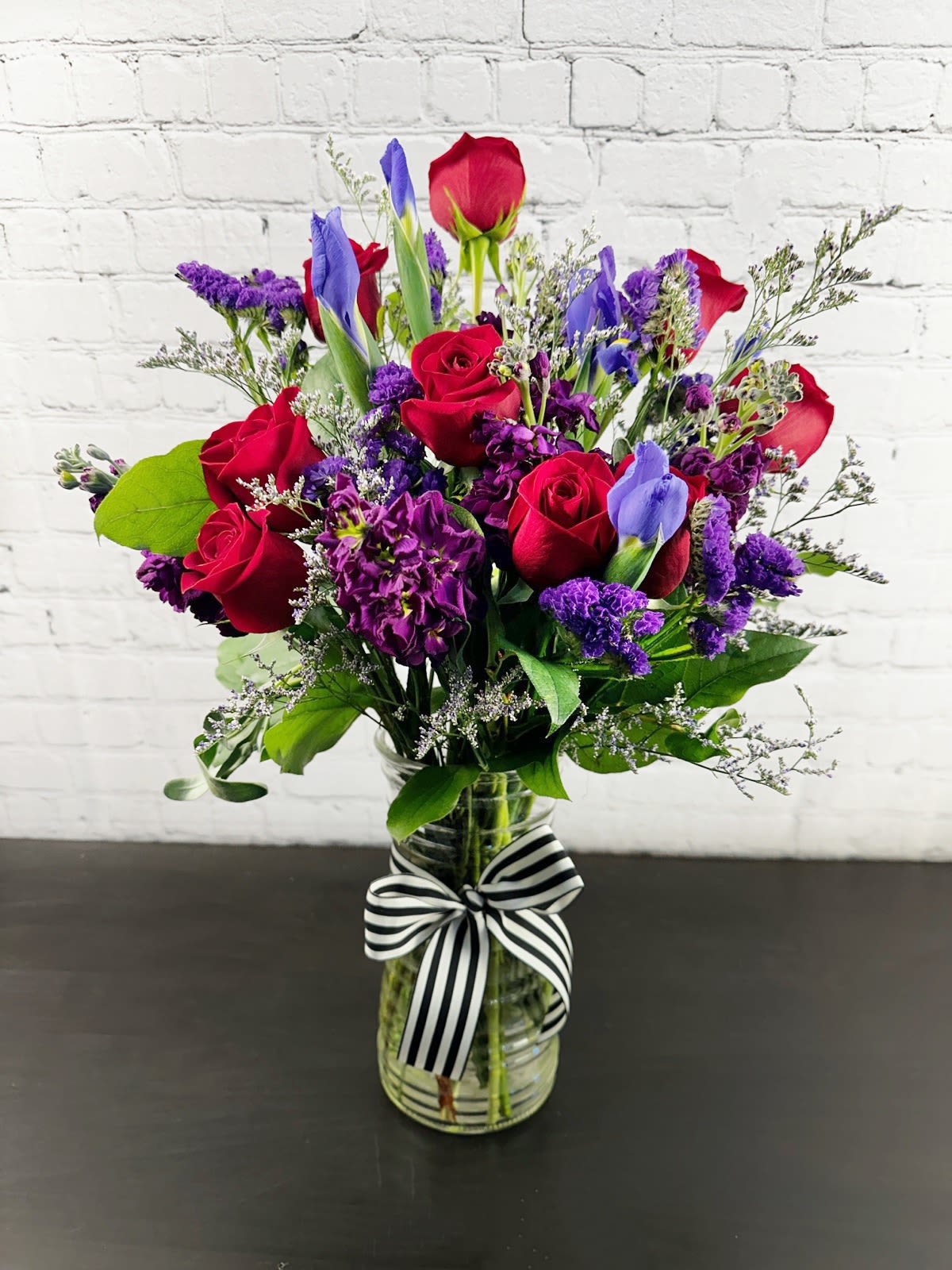 Deeply Loved - Bright and Vibrant jewel tones of red and deep purple set this arrangement apart from the others. Includes roses, irises, stock, and larkspur. Stands approximately 17" tall and 14" wide.   Please Note: Standard Delivery for this product occurs in the afternoon, between 2:30pm and 7pm. If you need it by a certain time, there is an option to select a timed window for scheduled deliveries.