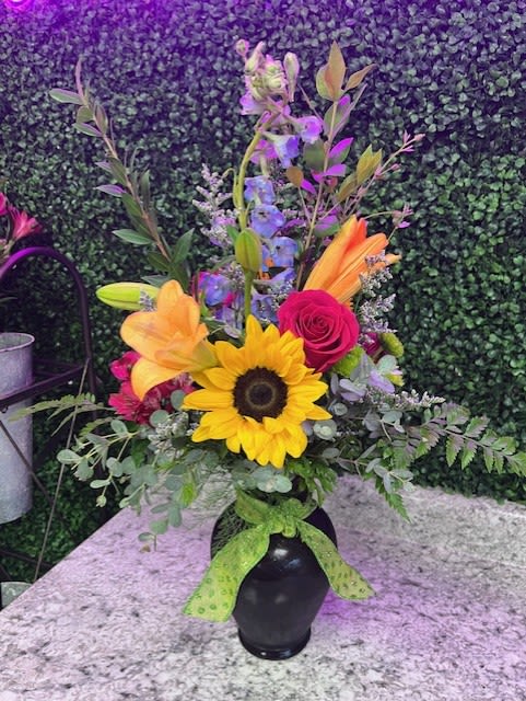 Tall mixed bouquet with sunflower, lilies, rose, and blue delphinium in a black vase with green bow.