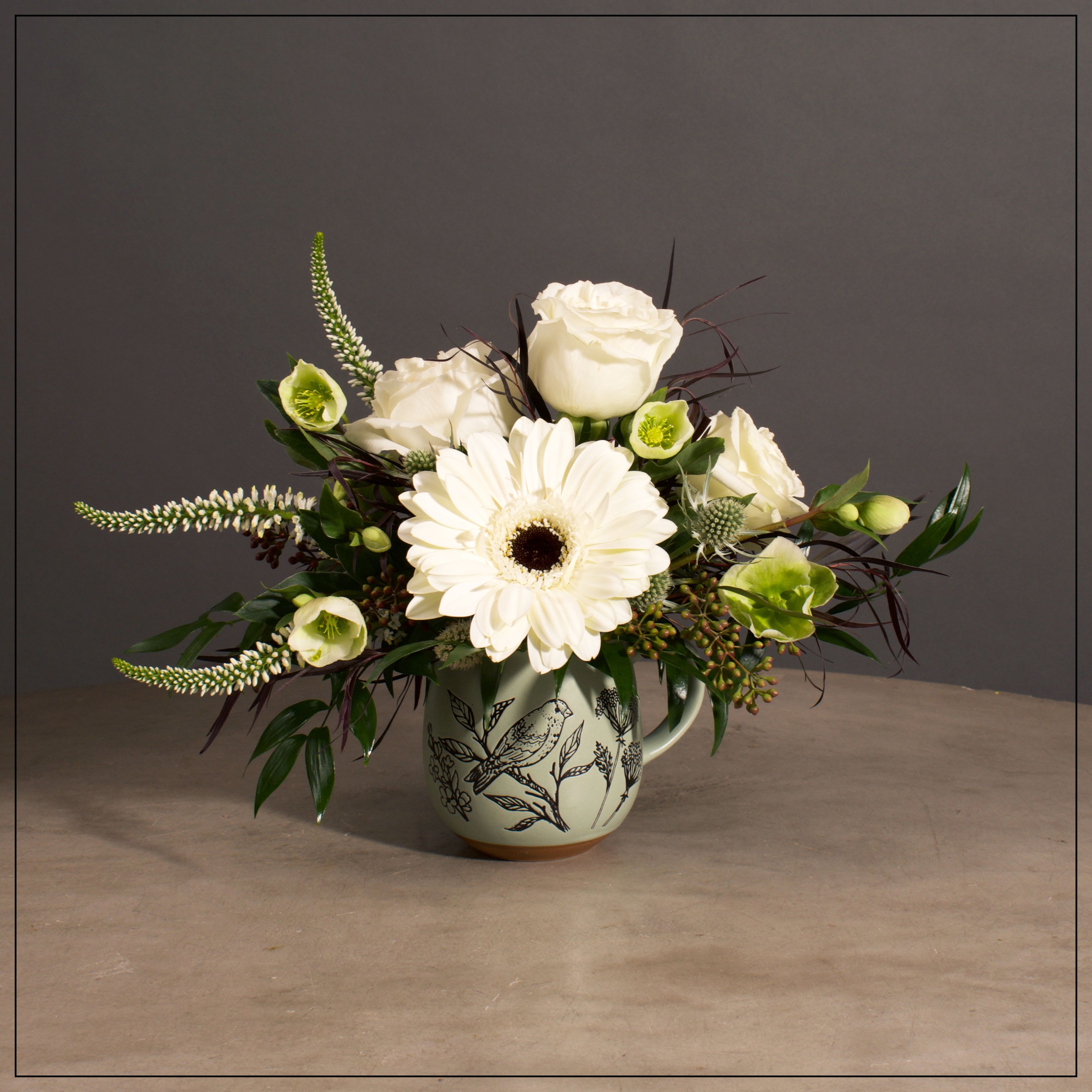 Bird Song - Gerber Daisies, roses, Veronica, Hellebores and Mums with Mixed Greens in a Ceramic Mug.  Approximately 8" H x 10" W