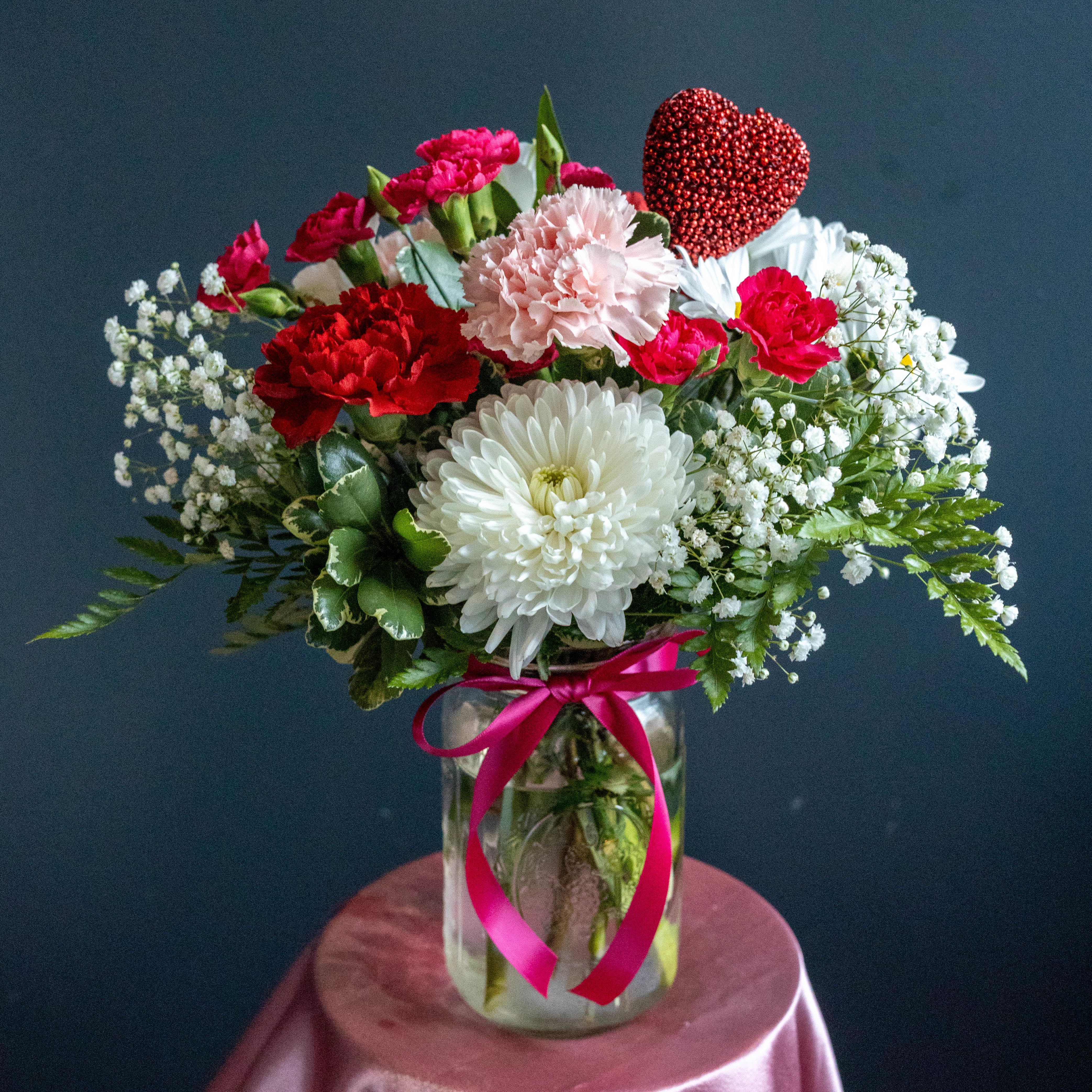 Sweet Lil Lollipop - Charming, cheerful, and full of love the Sweet Lil Lollipop is the perfect Valentine to brighten someone’s day. This delightful arrangement features a playful mix of soft pink and rich red carnations, crisp white mums, and delicate baby’s breath, all accented with fresh greenery for a light, airy feel.  A sparkling red heart pick adds a fun Valentine’s touch, while the clear Ball mason jar tied with a hot pink ribbon keeps the look sweet and simple. Designed to be compact yet full of personality, this arrangement is ideal for desks, side tables, or anywhere a pop of love is needed.  Cute, colorful, and irresistibly sweet, this one’s sure to make hearts smile. ️