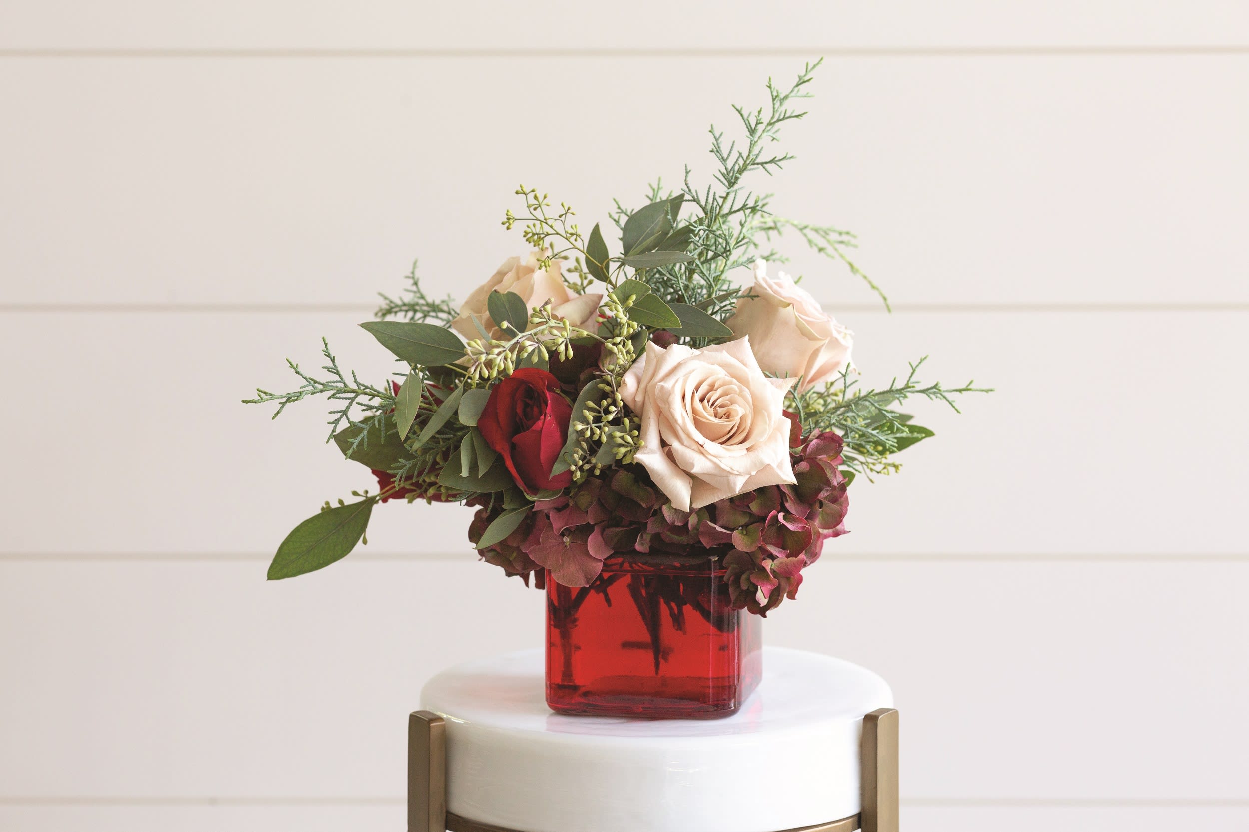 Crimson Poise – Petite Romantic Vase Arrangement - Crimson Poise is a refined expression of romance, designed with contrast, depth, and intentional elegance. Arranged in a rich red square glass vase, this petite composition features soft blush roses paired with a single deep red rose, grounded by lush seasonal greenery and subtle textural blooms for a balanced, garden-inspired silhouette.  This arrangement blends classic Valentine’s tones with a modern, restrained sensibility romantic without excess, expressive without overpowering. Its compact scale makes it ideal for intimate settings such as bedside tables, desks, or small dining moments where a thoughtful gesture speaks volumes.  Each Crimson Poise arrangement is crafted by our expert florists using premium seasonal blooms, ensuring beauty, freshness, and a polished finish in every detail.  Size Details: Compact, low-profile design  Petite square glass vase| Ideal for small spaces and refined gifting  Seasonal Notes Flower varieties may vary based on availability. The red, blush, and greenery palette, and the overall romantic aesthetic will always be maintained.  Ideal For: Valentine’s Day gestures, anniversaries, romantic thank-yous, or elegant everyday gifting.