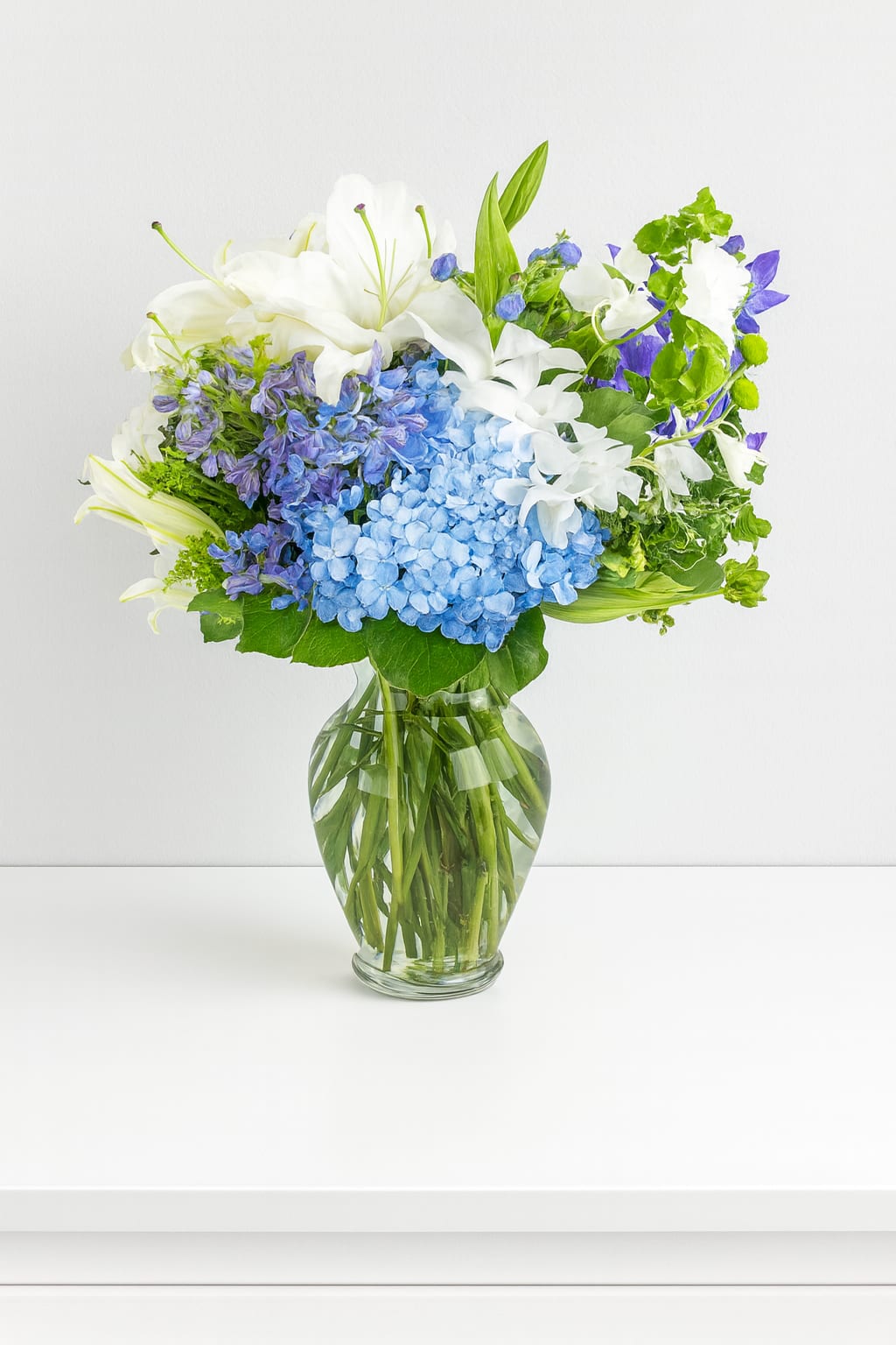 Breezy by the Sea by BloomNation™ - This arrangement contains delphinium, bells of ireland, asiatic lilies, hydrangea, blue iris, mokara orchids, and other seasonal blooms. It is a great gift for a birthday, get well, for wishing someone a fresh start. APPROXIMATE DIMENSIONS: 15" D x 18" H