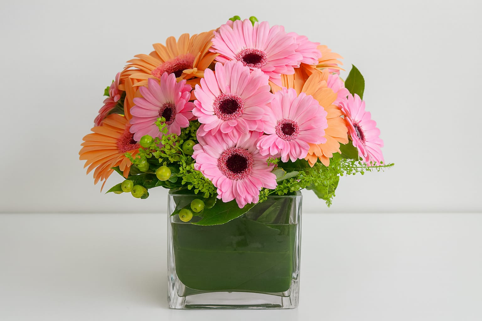 Bright As Daytime - Assorted pink and peach Gerbera Daisies designed with an assortment of novelty greens in a leaf wrapped 5 inch glass cube.  Will brighten someones room or desk, as well as their spirits.  Available in most color palettes; specify any preference  in 'special instruction' box.