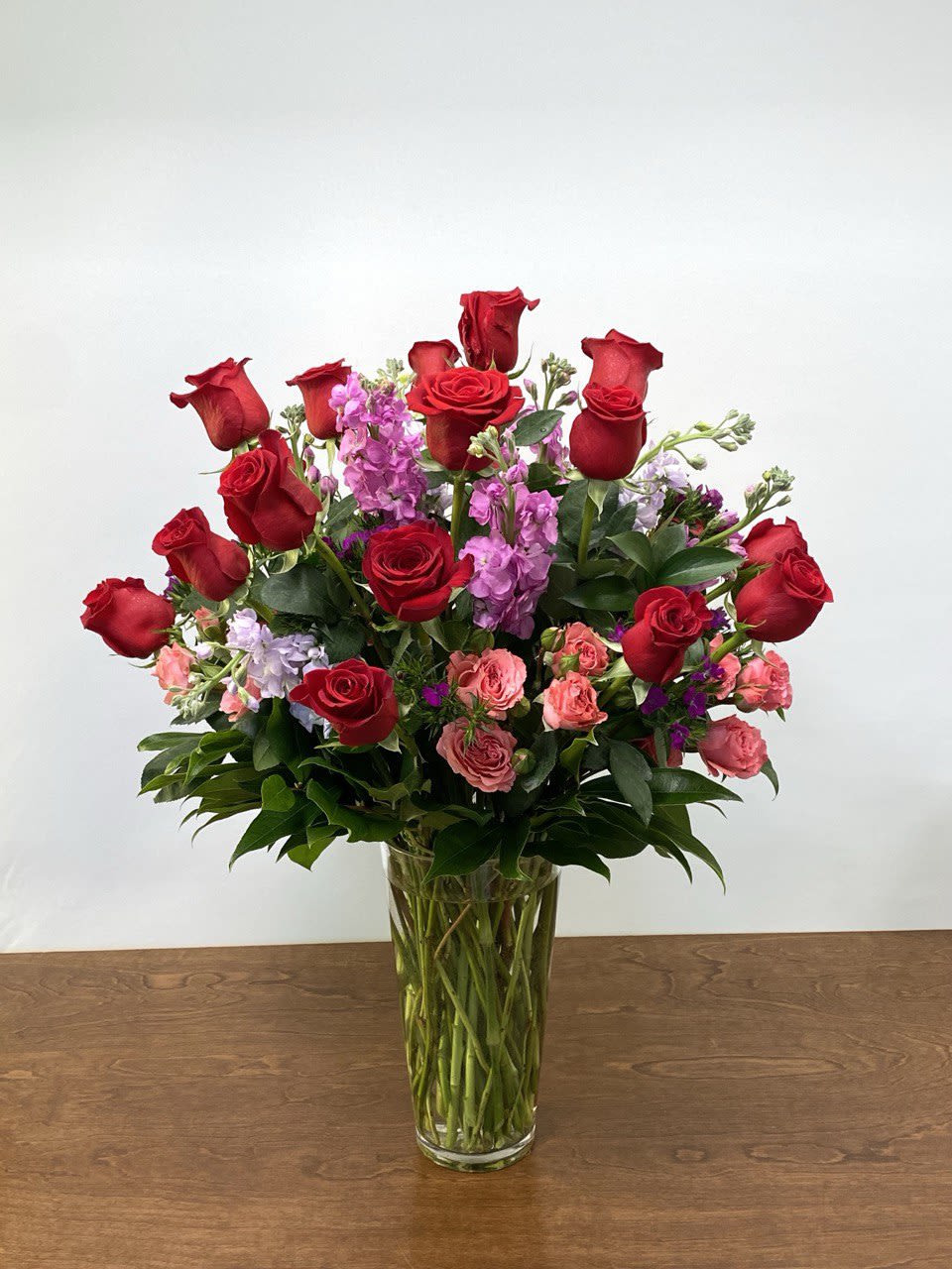 Rose Garden - A vibrant and garden-inspired arrangement showcasing premium red roses accented with soft pink spray roses, seasonal lavender blooms, and lush greenery. Designed tall and full in a clear glass vase, this arrangement offers beautiful height, movement, and rich texture. Elegant yet cheerful, it’s a striking choice for Valentine's Day, birthdays, anniversaries, celebrations, or making a bold statement in any space.
