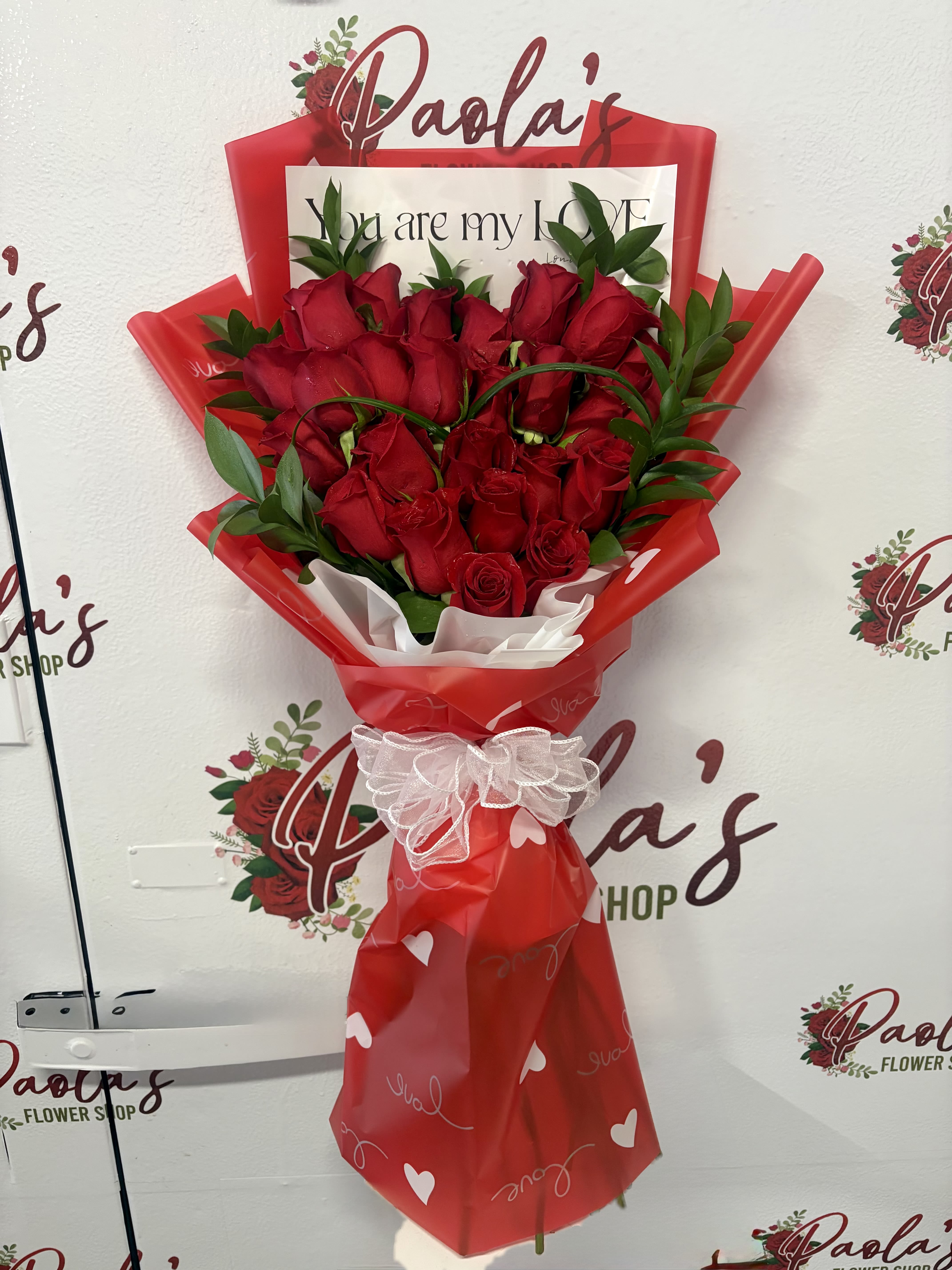 Hidden Heart Rose Bouquet - Say “I love you” in the most romantic way with our Hidden Heart Rose Bouquet. This stunning arrangement features premium red roses expertly designed to form a softly obscured heart shape within the bouquet, creating a beautiful and meaningful surprise as the flowers are admired from every angle.  Wrapped in elegant love themed paper and finished with a delicate ribbon accent, this bouquet is perfect for anniversaries, Valentine’s Day, or simply expressing heartfelt love. The lush greenery enhances the rich red roses, adding depth, texture, and a timeless romantic feel.  *Does NOT include vase*