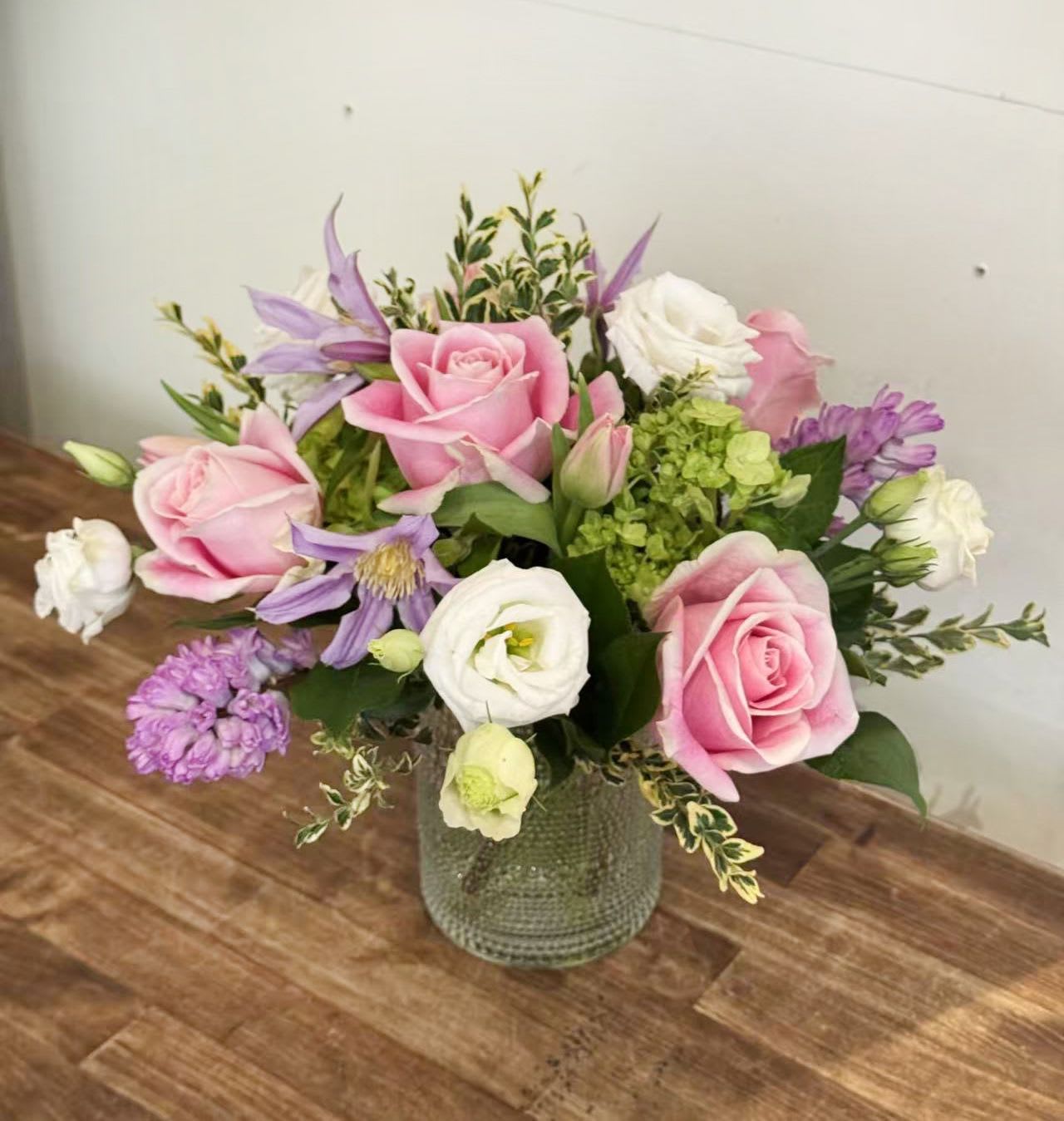 Petal Whisper - Soft, romantic, and effortlessly elegant, this arrangement is a celebration of gentle color and natural beauty. Blush pink roses take center stage, paired with creamy white lisianthus, delicate purple clematis, and fresh green hydrangea accents. Thoughtfully layered with seasonal greenery, the design feels light, garden-inspired, and beautifully balanced.  Arranged in a textured glass vase, this piece is perfect for birthdays, thank-you gifts, celebrations, or simply bringing a touch of calm and charm into any space—home or office.  Details: 	•	Features pink roses, white lisianthus, purple clematis, green hydrangea, and seasonal foliage 	•	Soft pastel palette with a fresh garden feel 	•	Designed in a modern textured glass vase 	•	Hand-designed for a natural, airy look  Please note: Flower varieties may vary slightly based on seasonal availability, while maintaining the overall color palette and style.