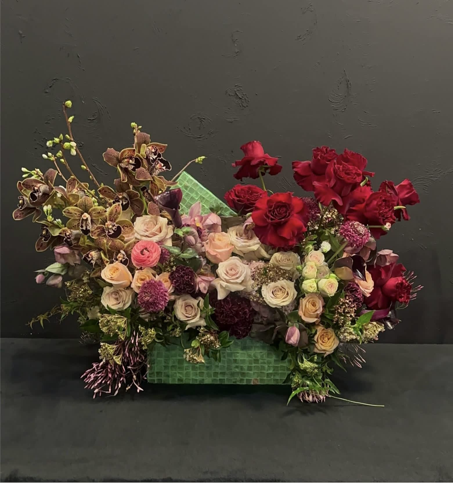 Enchanting Elegance - Enchanting Elegance embodies the epitome of floral sophistication. At its heart, pristine cymbidium orchids stand tall, exuding an aura of timeless grace. Surrounding them are delicate peach and red roses, in a symphony of passion and tenderness. Interspersed among them. This arrangement, with its harmonious blend of colors and textures, evokes a sense of romance and enchantment, making it a captivating centerpiece for any occasion.