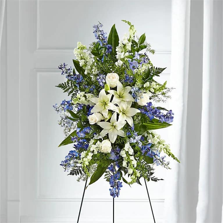 Faithful Friend Standing Spray - A collection of bold blue flowers shares a tribute that words cannot express. Designed with lilies, roses, delphinium and snapdragons our Faithful Friend Standing Spray is a grand gesture to express kindness and solace to a loved one.
