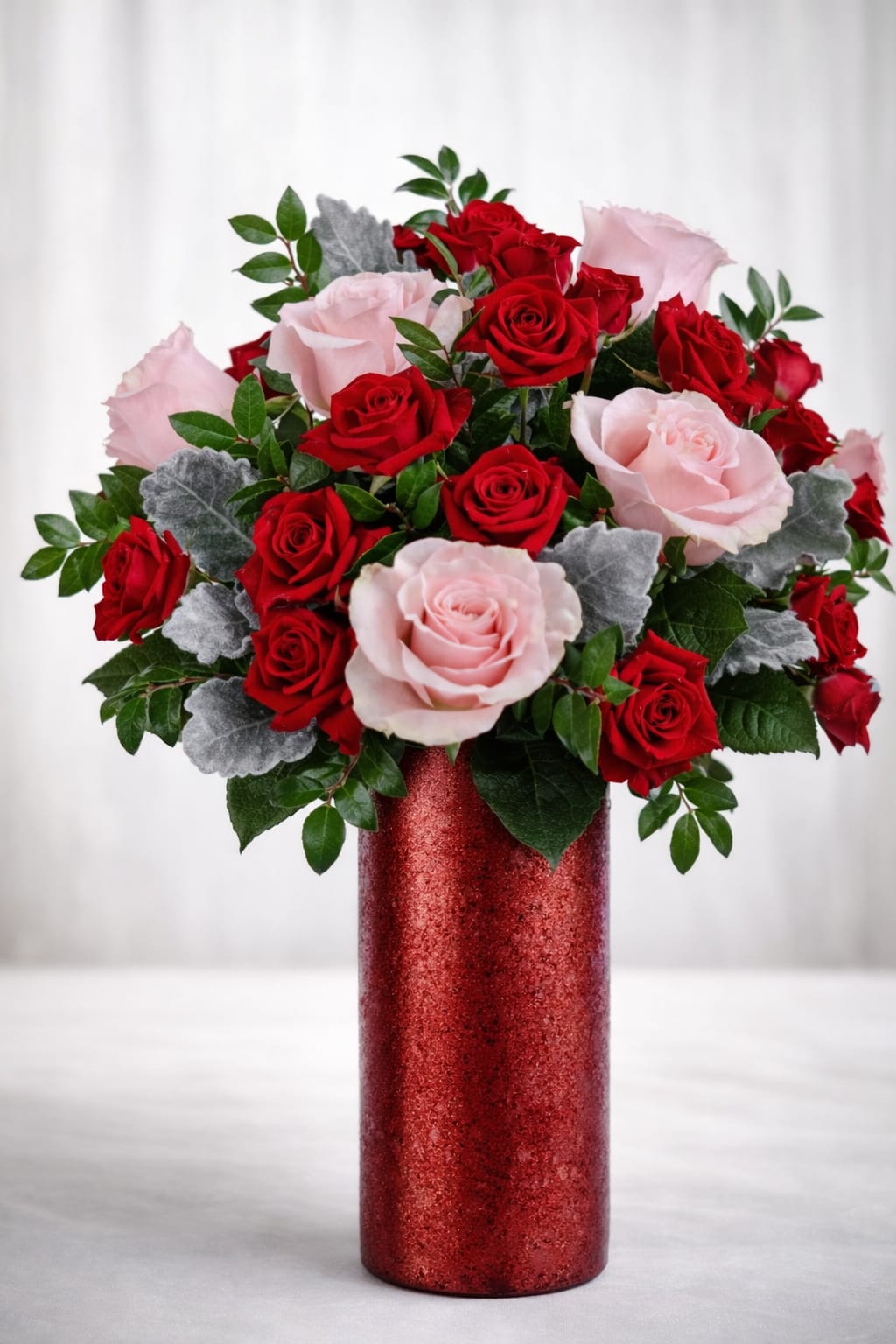 Blush &amp; scarlet elegance  -  A stunning mix of velvety red roses and soft blush pink roses, accented with textured silver-tone foliage and fresh greenery. Designed in a tall, shimmering red vase for a graceful, upscale presentation.