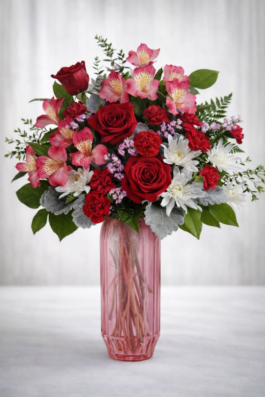 Sweet  romance garden  -  A lush mix of red roses, pink alstroemeria, white mums, red carnations, and soft accent flowers, finished with fresh greenery and dusty miller. Designed in a translucent blush glass vase for a light, elegant look