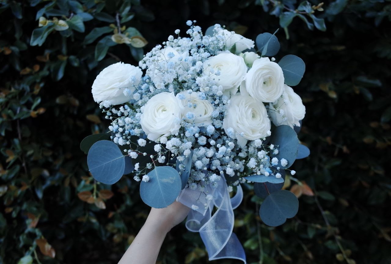 Whispers of Blue Bridal Bouquet - This hand-tied bouquet blends classic white ranunculus with delicate blue and white filler flowers and fresh eucalyptus for a naturally graceful look. Finished with a sheer blue ribbon, it’s a refined and romantic choice for a spring or garden-inspired wedding.