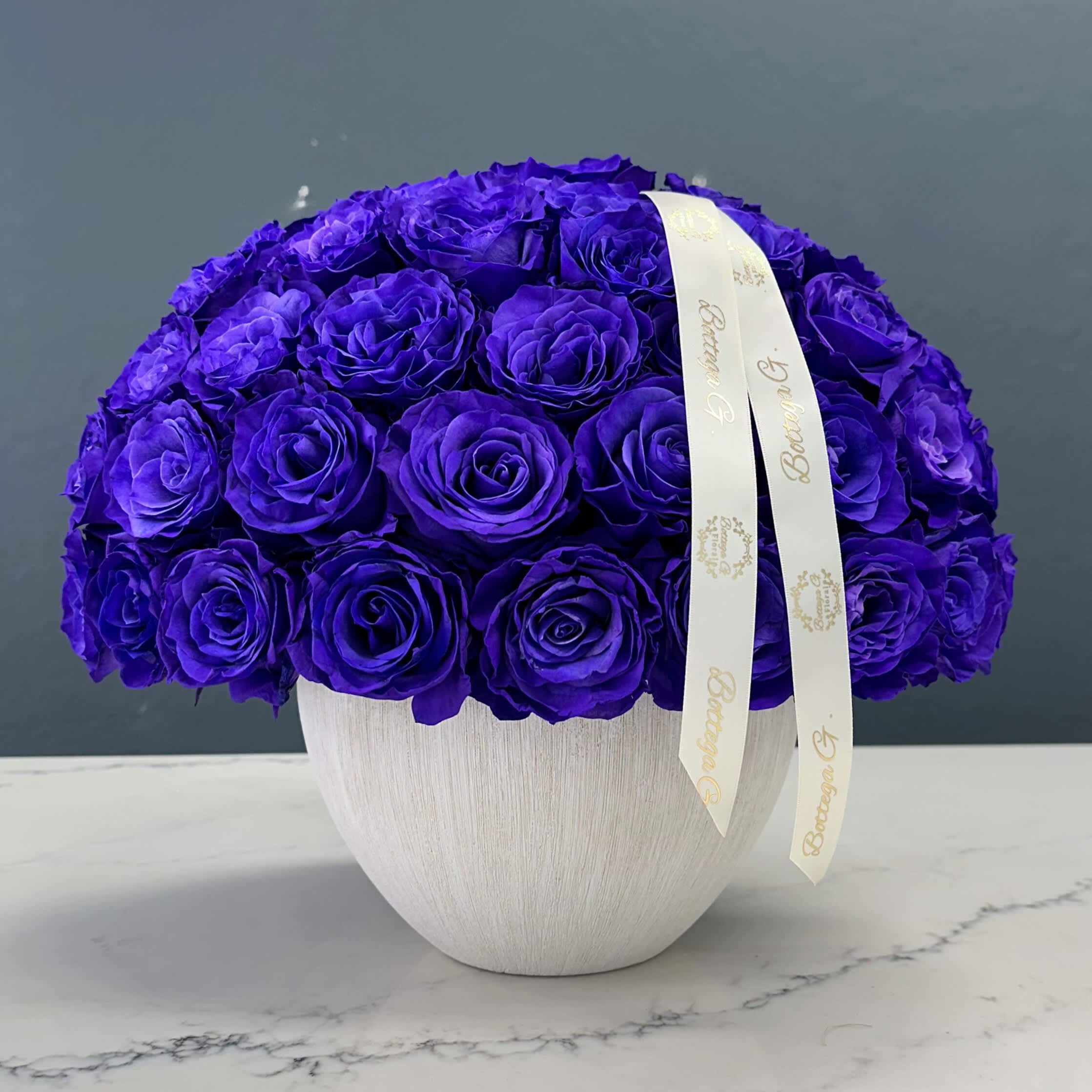 Grape Bliss - Imagine an exquisite arrangement of purple roses elegantly displayed in a charming rustic vase. The roses, in various shades of lavender and deep violet, exude a sense of mystery and sophistication. Each bloom is carefully selected for its velvety petals and lush, full appearance, creating a harmonious blend of color and texture.  The rustic vase, with its weathered finish and earthy tones, adds a touch of countryside charm to the display. Its natural imperfections and textured surface perfectly complement the rich hues of the roses, creating a striking contrast that highlights the flowers' beauty. The vase might be crafted from aged ceramic or distressed wood, featuring subtle patterns or engravings that enhance its antique appeal.  Accompanying the roses are sprigs of fresh greenery and delicate filler flowers, such as baby's breath or wax flowers, adding layers of depth and a hint of wild elegance. This arrangement, with its blend of vibrant blooms and rustic charm, evokes a sense of timeless beauty, making it a perfect centerpiece for any occasion, from a cozy home gathering to a sophisticated event.